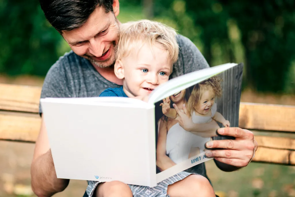 Father and young child sitting on a bench, joyfully reading a photo book together outdoors.