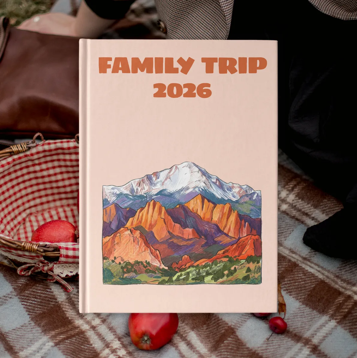 A book titled "Family Trip 2026" featuring a colorful mountain illustration, placed on a plaid blanket next to a picnic basket and apples.