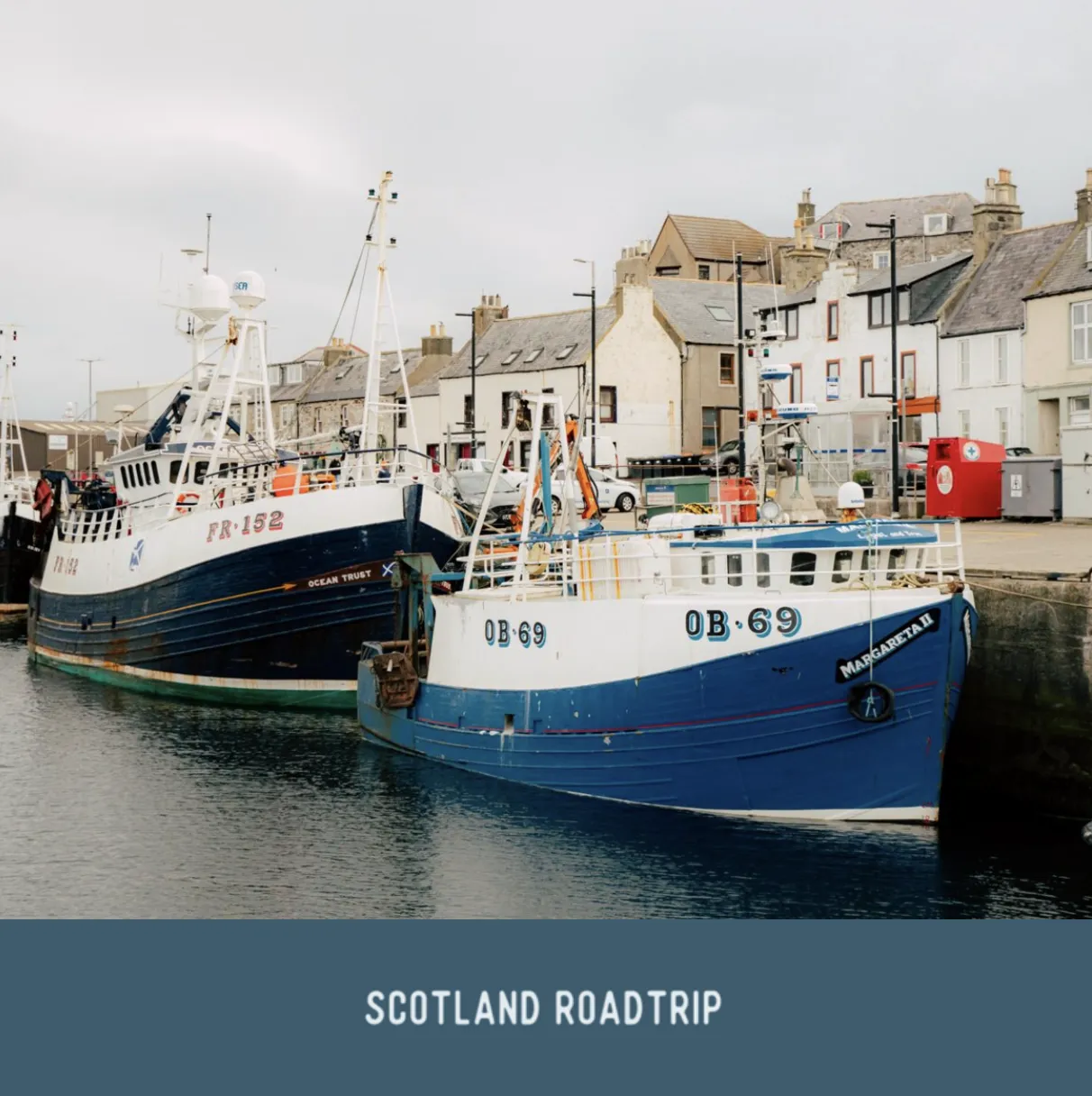Zwei blaue Fischerboote liegen in einem Hafen vor einer Reihe von Steinhäusern. Text: „Scotland Roadtrip“ unter dem Bild.