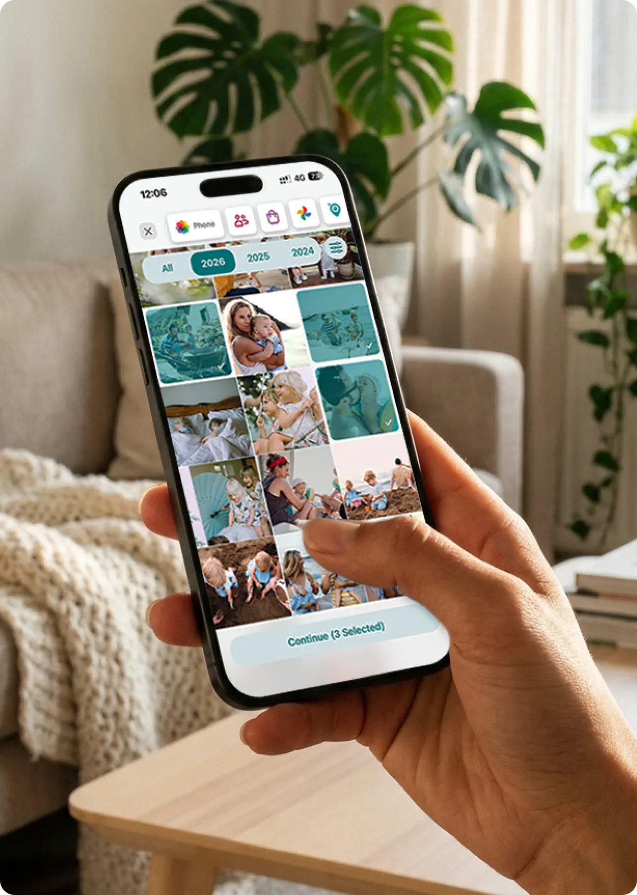 A hand holds a smartphone displaying a photo gallery with family pictures, inside a cozy living room with plants and a sofa in the background.