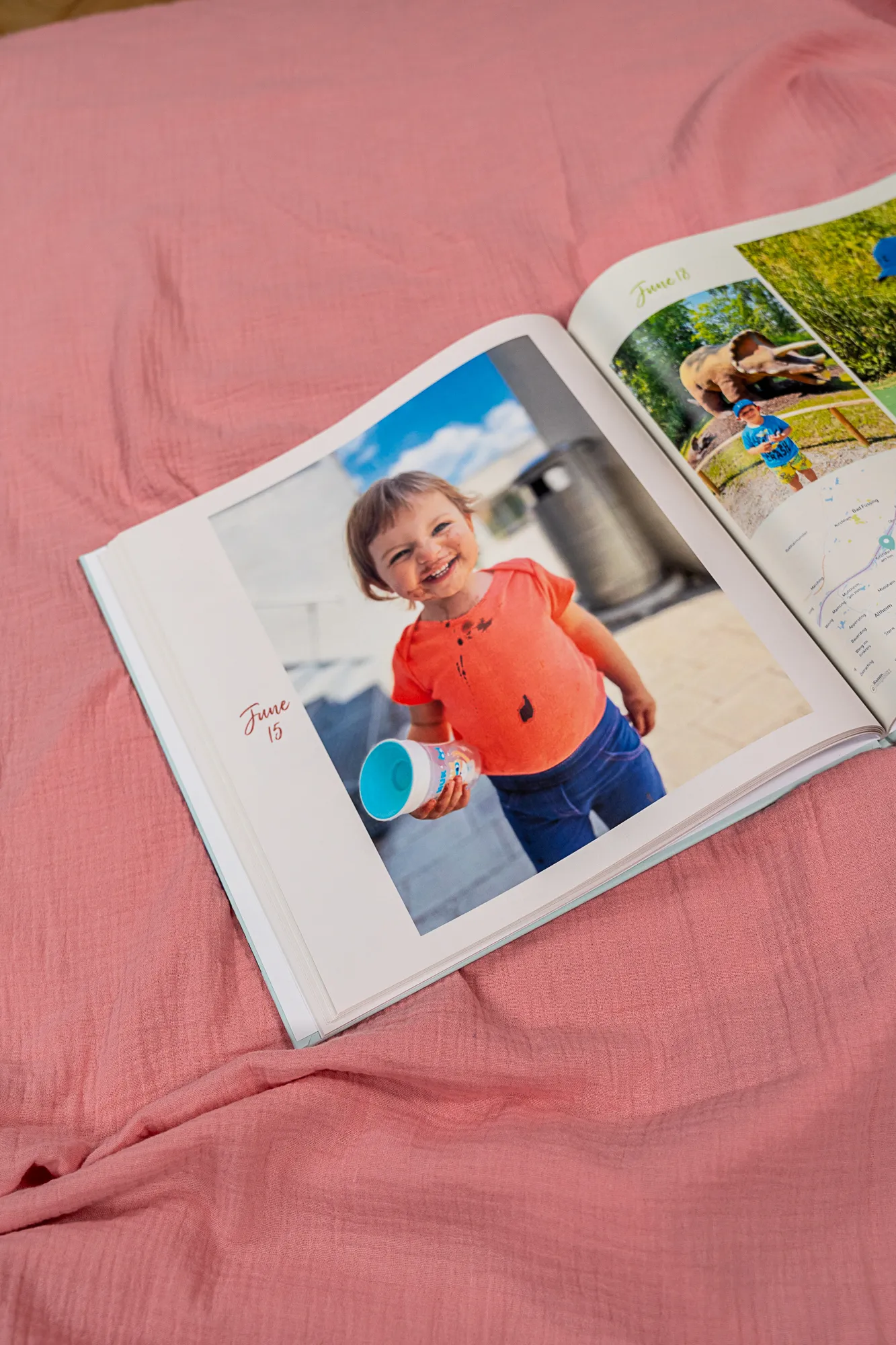 Ein offenes Fotobuch auf einer rosa Decke, auf dem ein lächelndes Kind zu sehen ist, das einen Behälter in der Hand hält; auf der Seite steht das Datum „15. Juni“.