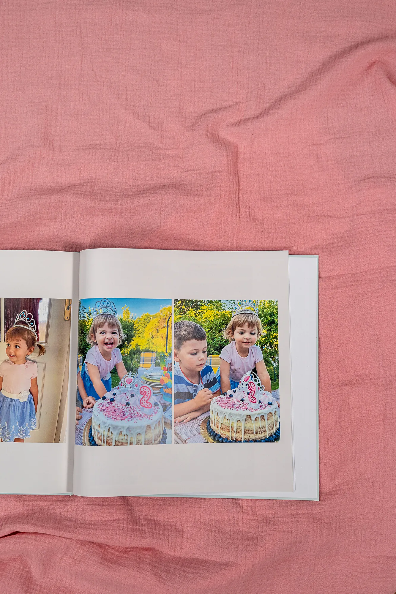 Geöffnetes Fotobuch auf rosa Stoff, das ein Kind mit einer Geburtstagstorte und einer Kerze in Form der Zahl 2 zeigt.