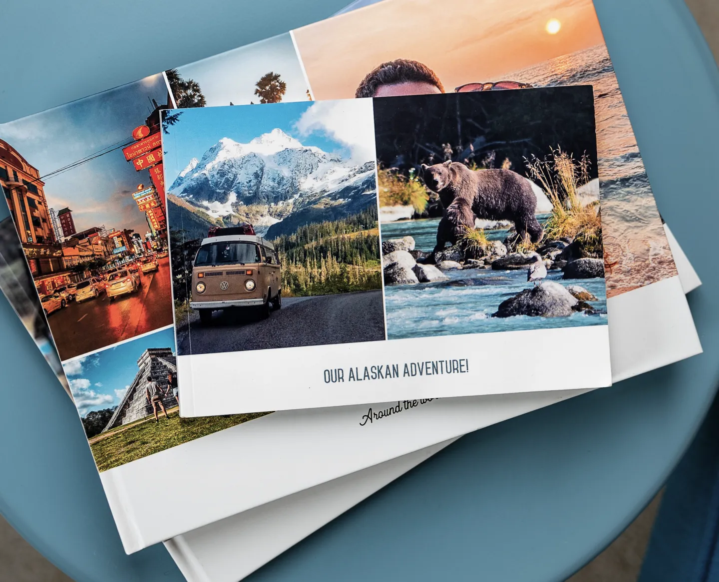 A photo book titled "Our Alaskan Adventure!" featuring images of a van in the mountains and a bear by a river, on a stack of similar books.