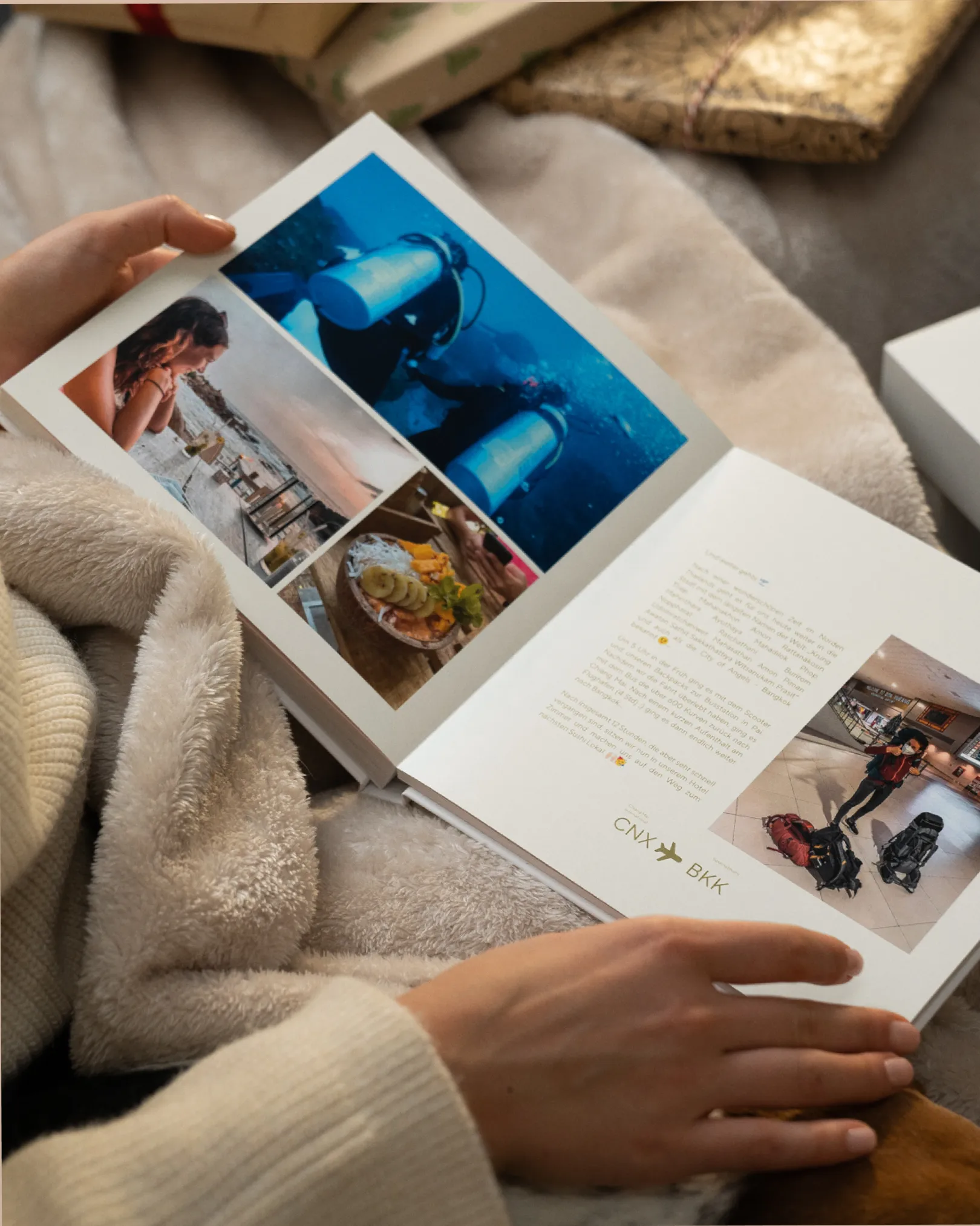 Person reading a photo book featuring travel images, including scuba diving and dining, while wrapped in a cozy blanket.