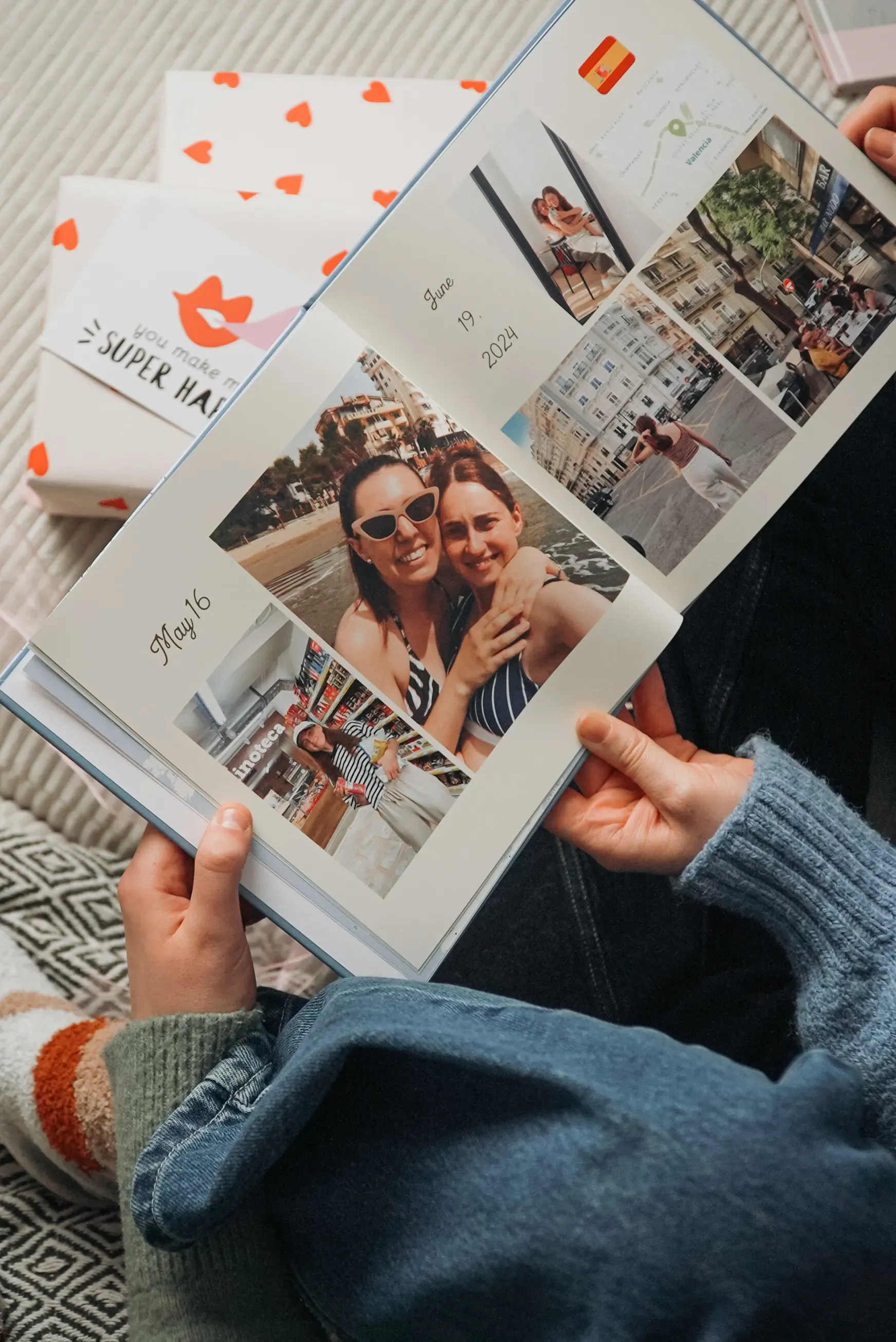 Person holding an open photo book featuring travel photos and handwritten notes. Gift boxes in the background.