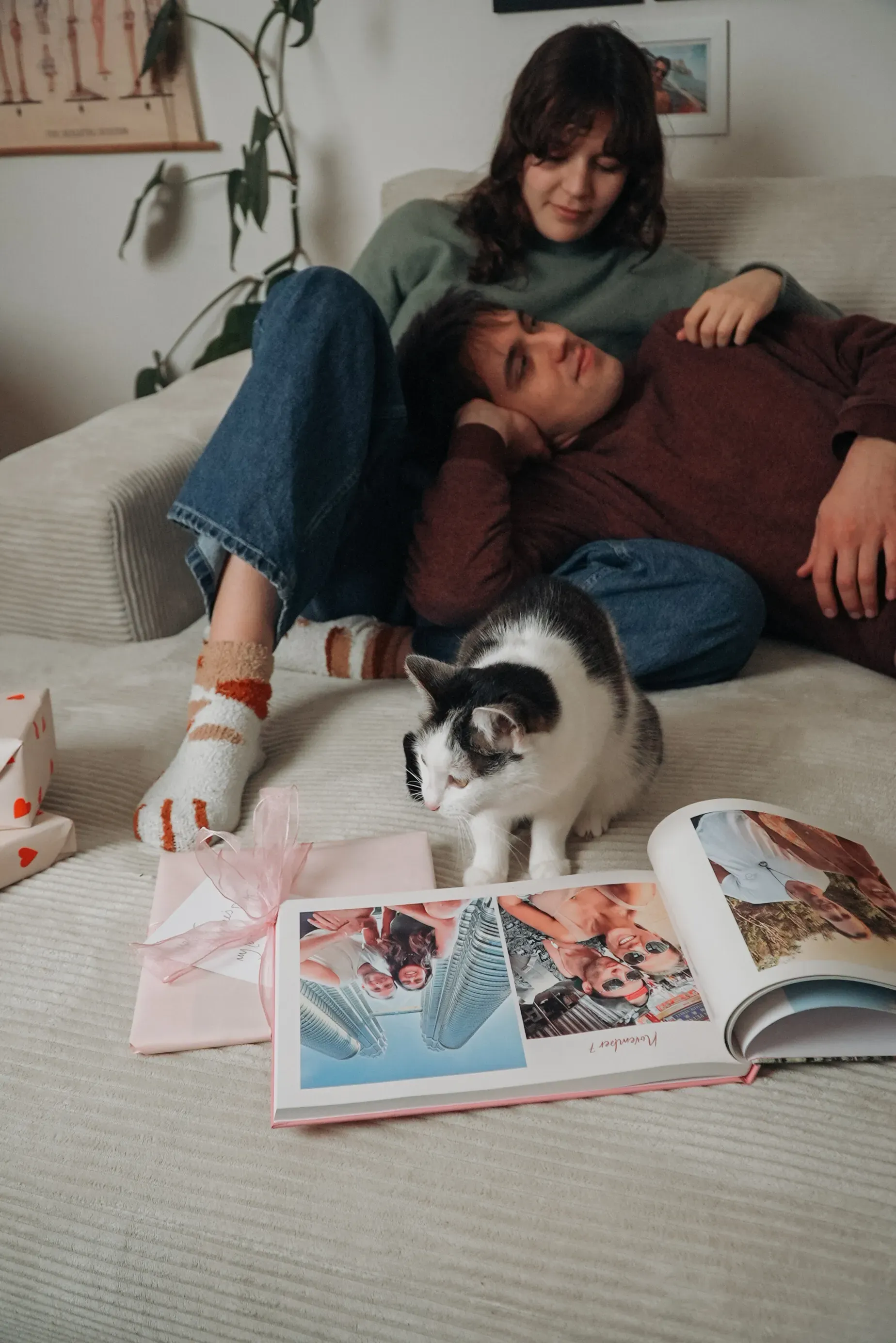 A couple relaxing on a sofa with a cat, a photo book, and a gift nearby. The woman is resting her head on the man's chest.
