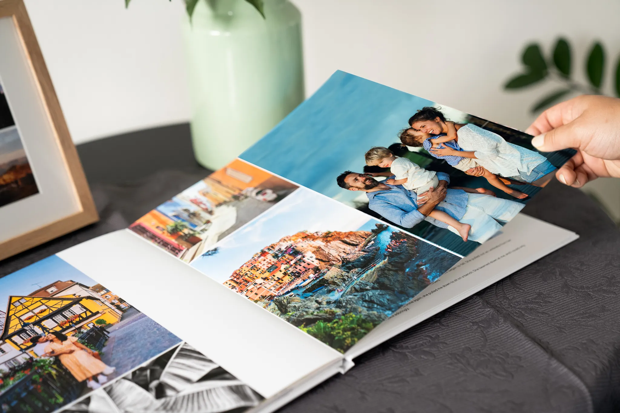 A hand flips through a photo book with colorful travel and family pictures on a table, next to a green vase and framed photo.