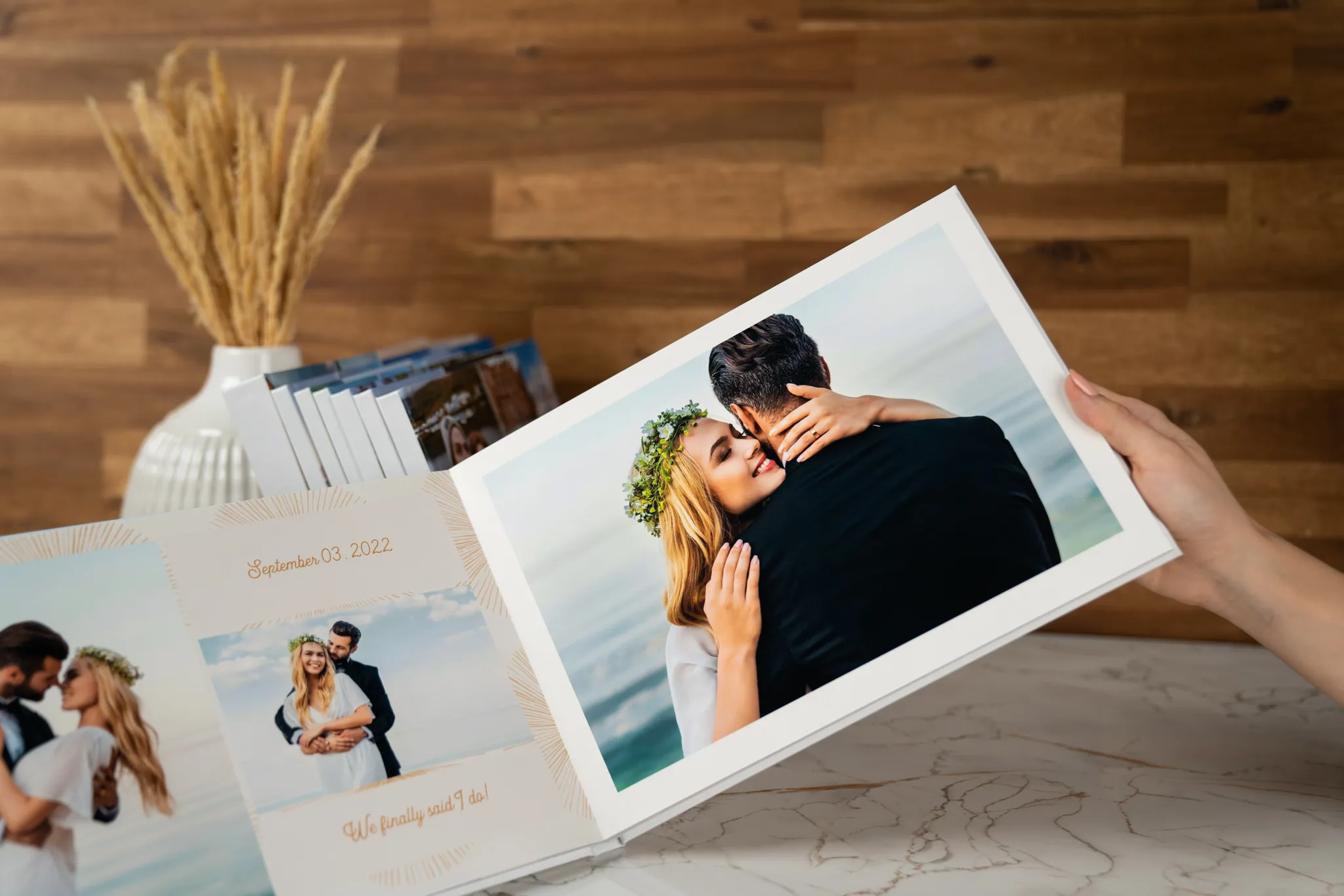 Eine Person hält ein aufgeschlagenes Hochzeitsfotobuch in der Hand, auf dem ein Paar zu sehen ist, das sich am Strand umarmt, im Hintergrund sind dekorative Vasen und eine Holzwand zu erkennen.
