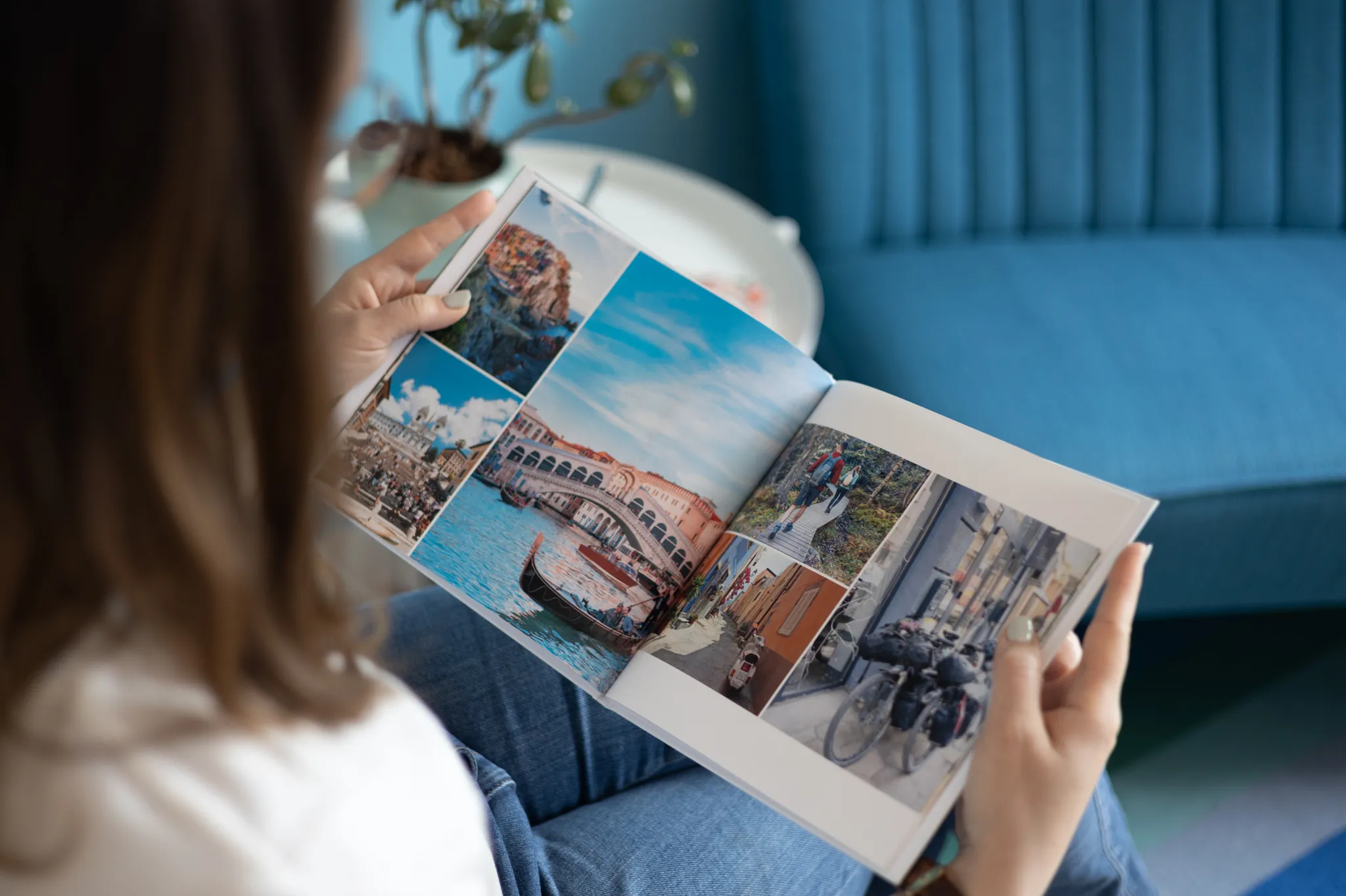 A person looks at a photo book featuring images of a scenic canal, a bridge, a motorcycle, and a street scene while sitting on a blue sofa.