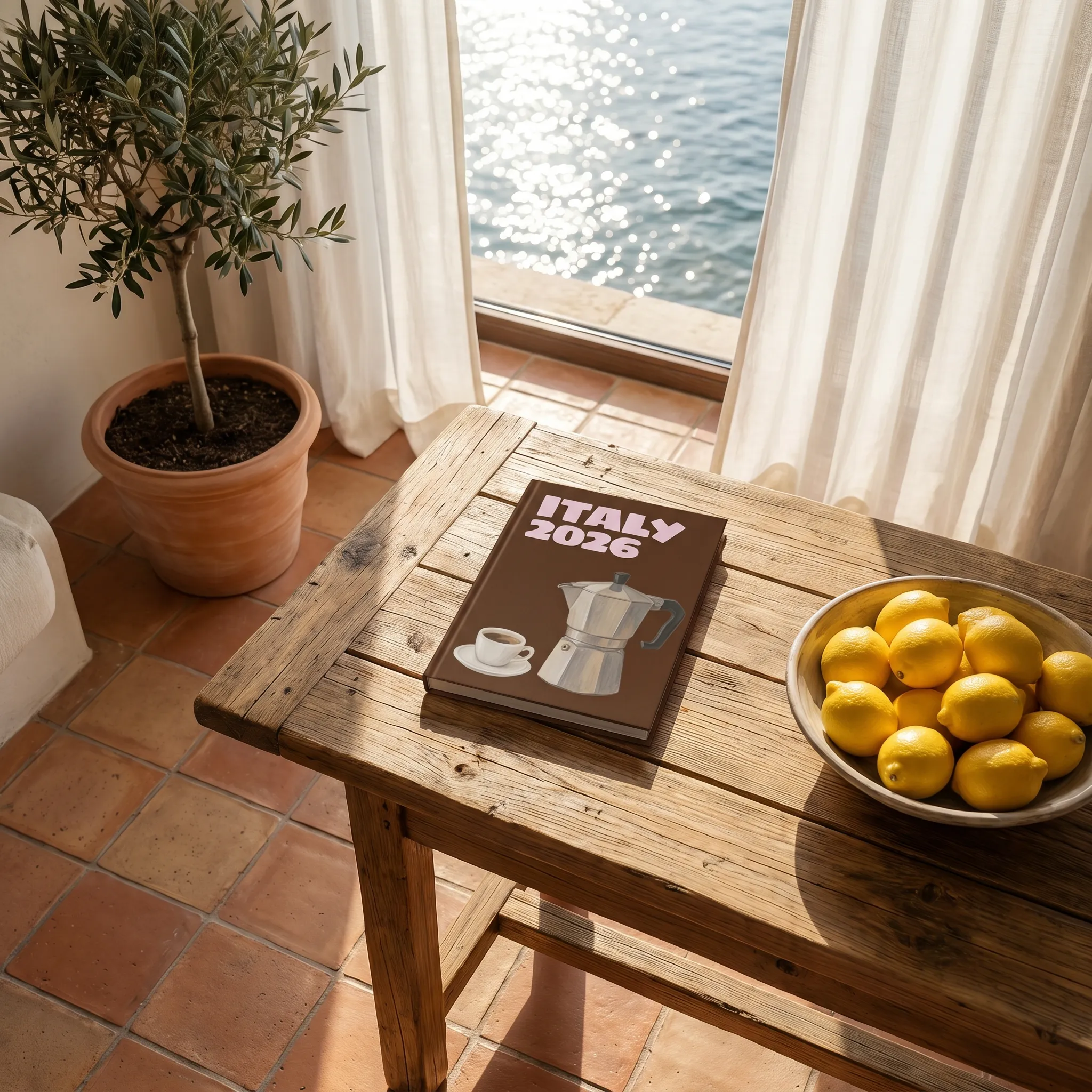 A rustic table with a photo book titled "Italy 2026," a plate of lemons, and an olive plant in a sunlit room overlooking water.