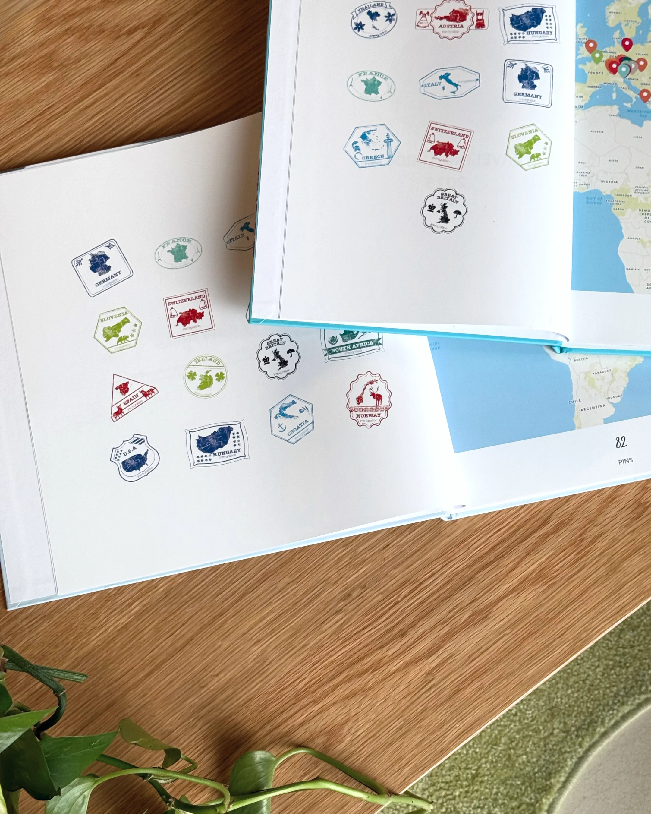 Two open travel photo books with colorful stamps and maps on a wooden surface, next to a green plant.