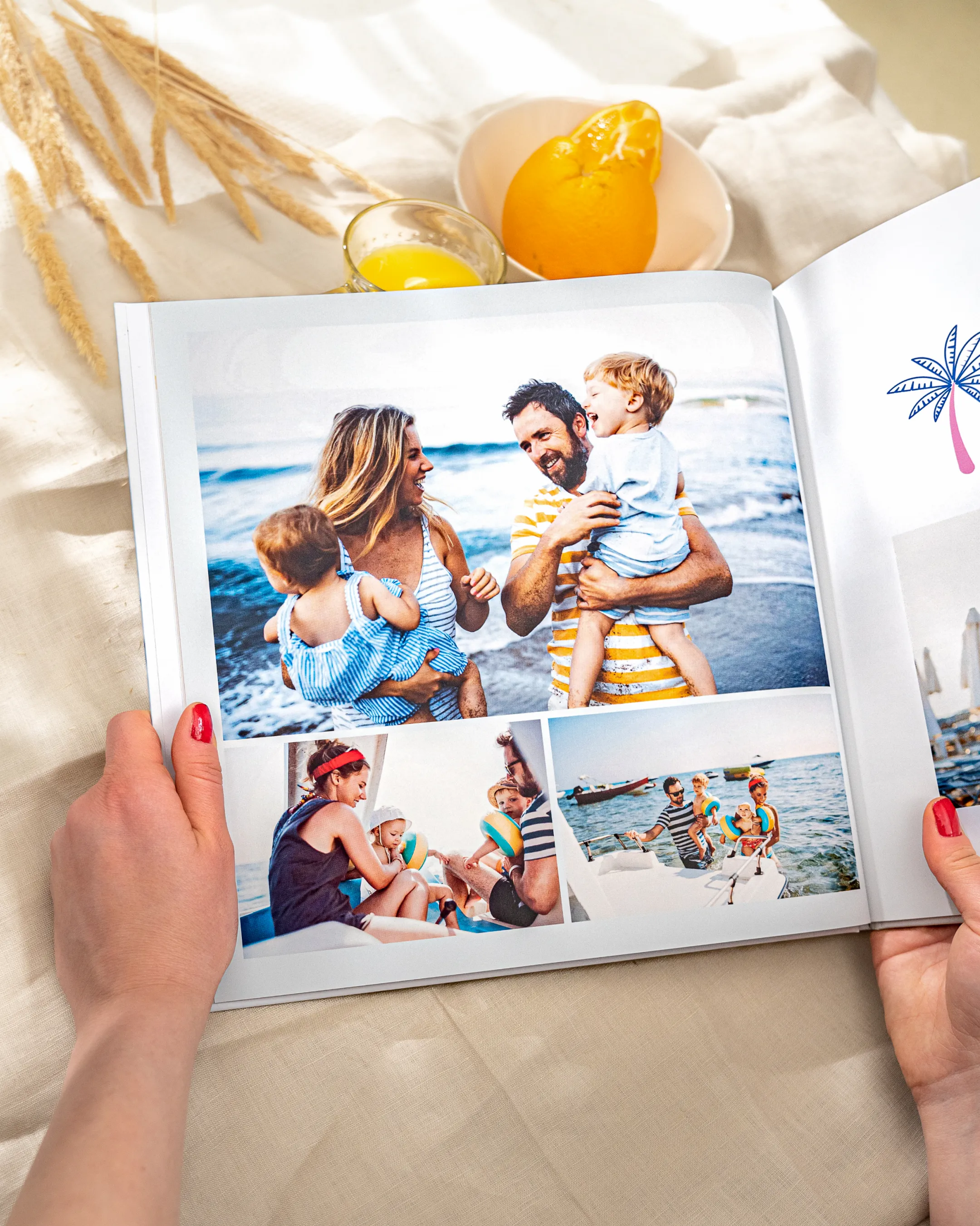 Person holding a photo album with beach family photos; nearby are a glass of juice and an orange on a tablecloth.