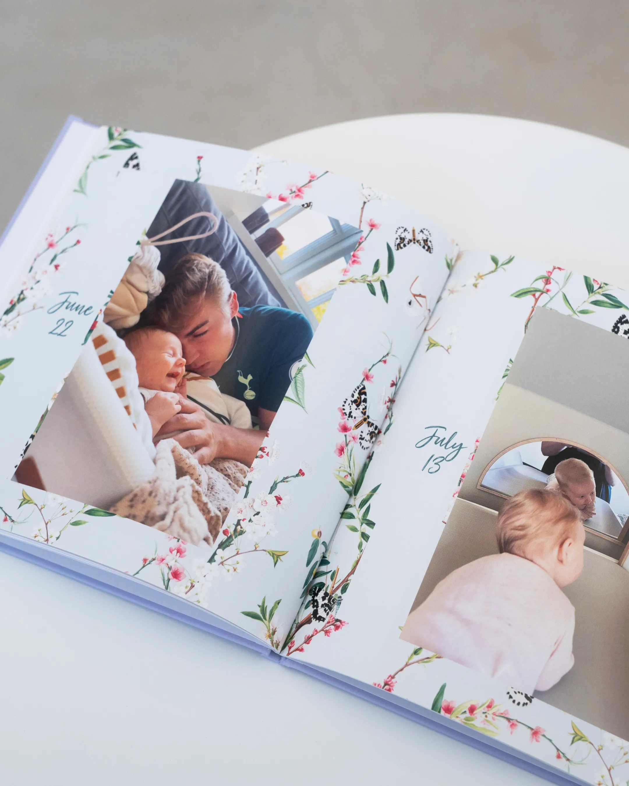 Open baby photo album with floral design, showing a man cuddling a baby on June 22 and a baby looking out a window on July 13.