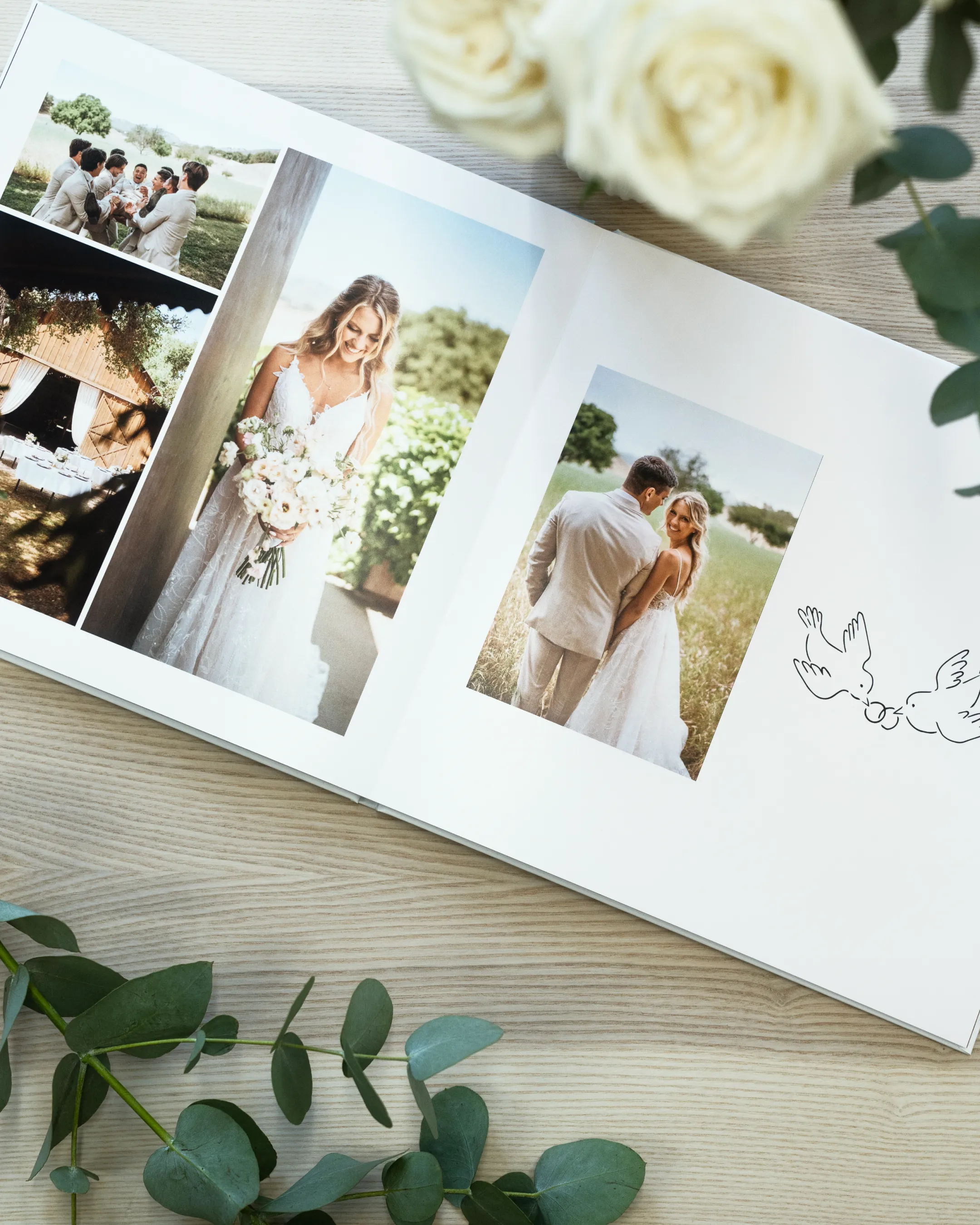 Open wedding photo book on a wooden table, surrounded by white roses and eucalyptus leaves.