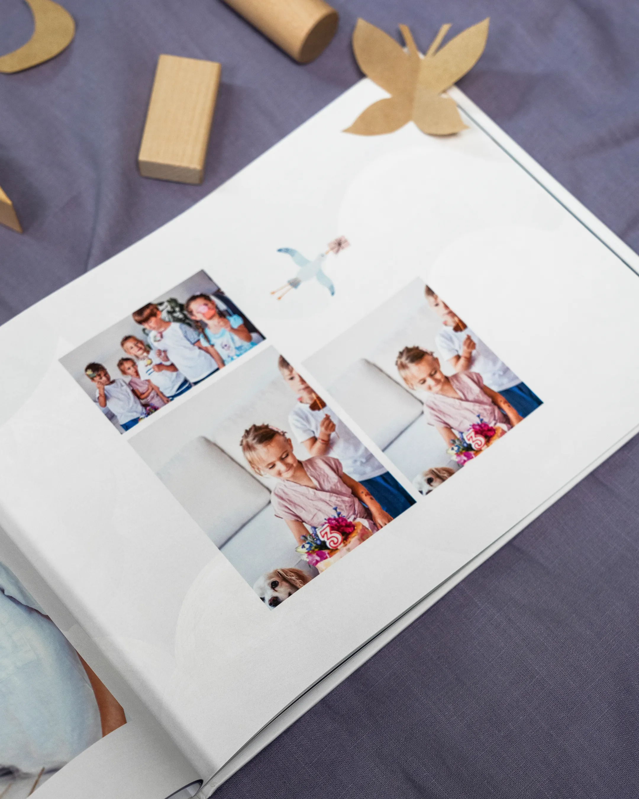 Open photo album on a gray fabric surface, featuring children and a dog in colorful, candid moments. Wooden blocks and a butterfly cutout nearby.
