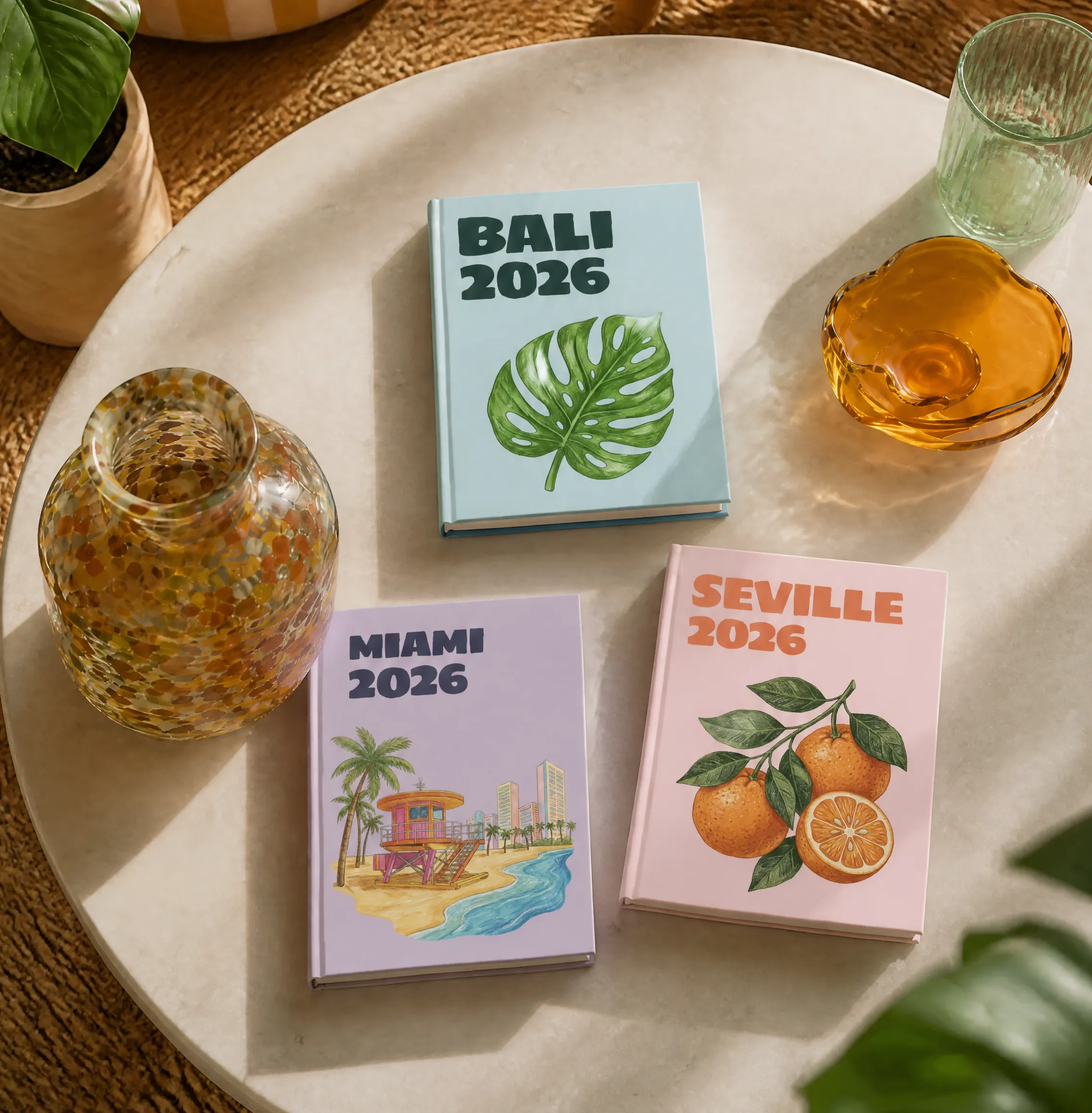 Three colorful 2026 travel journals on a table, labeled Bali, Miami, and Seville, with themed illustrations. Decorative vase and glass nearby.