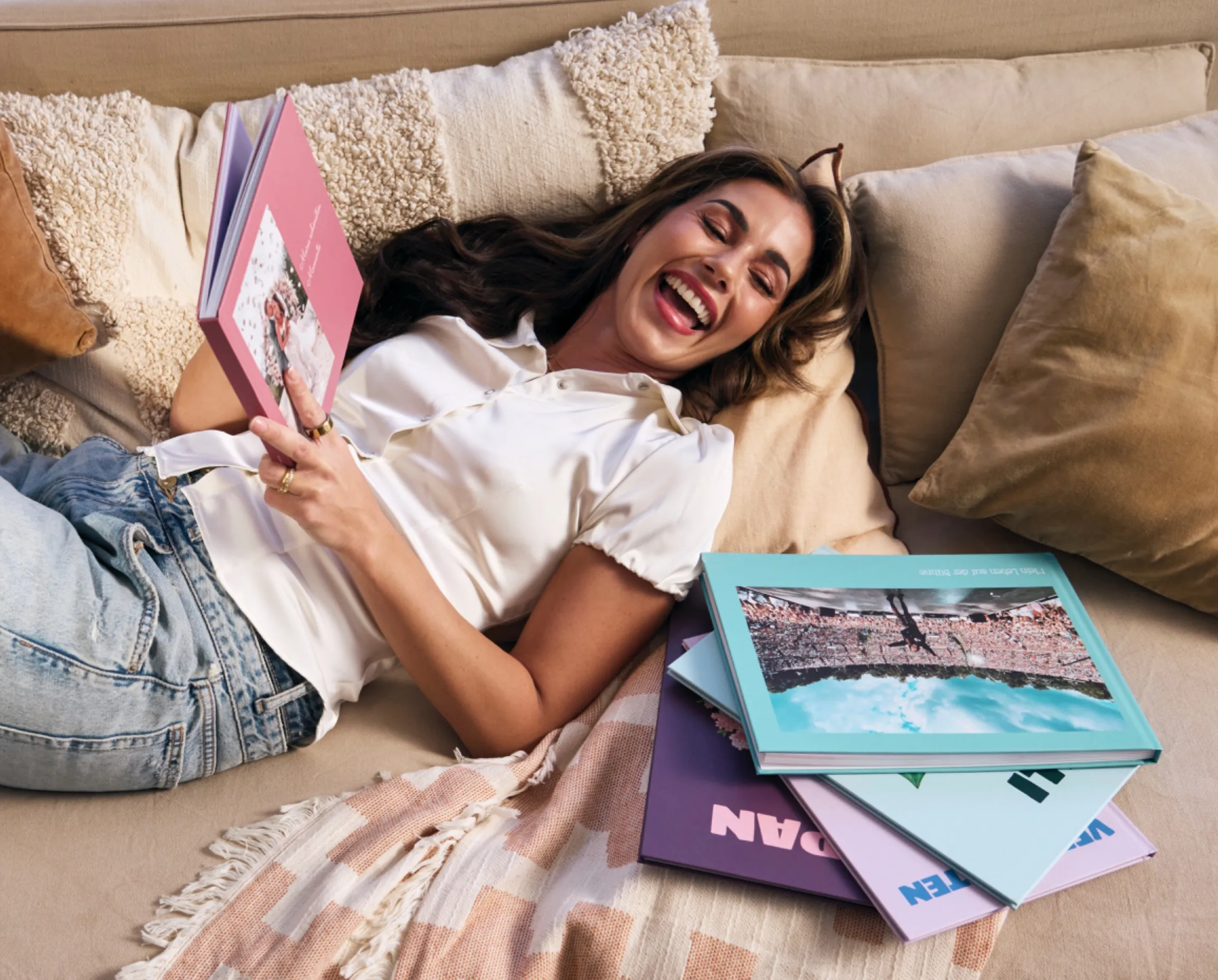 Sara Engels joyfully reading a Journi photo book on a cozy couch with pillows and more books nearby.
