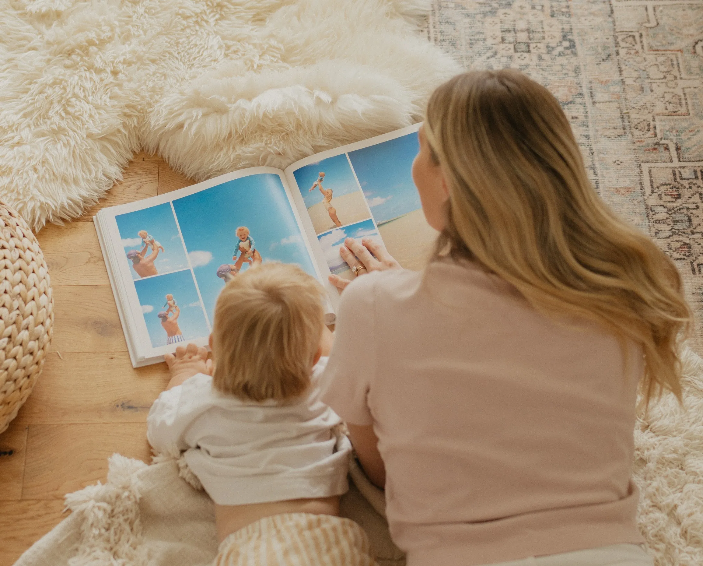 A woman and a child lie on a fluffy rug, looking at a photo book featuring beach scenes, creating a cozy and serene atmosphere.
