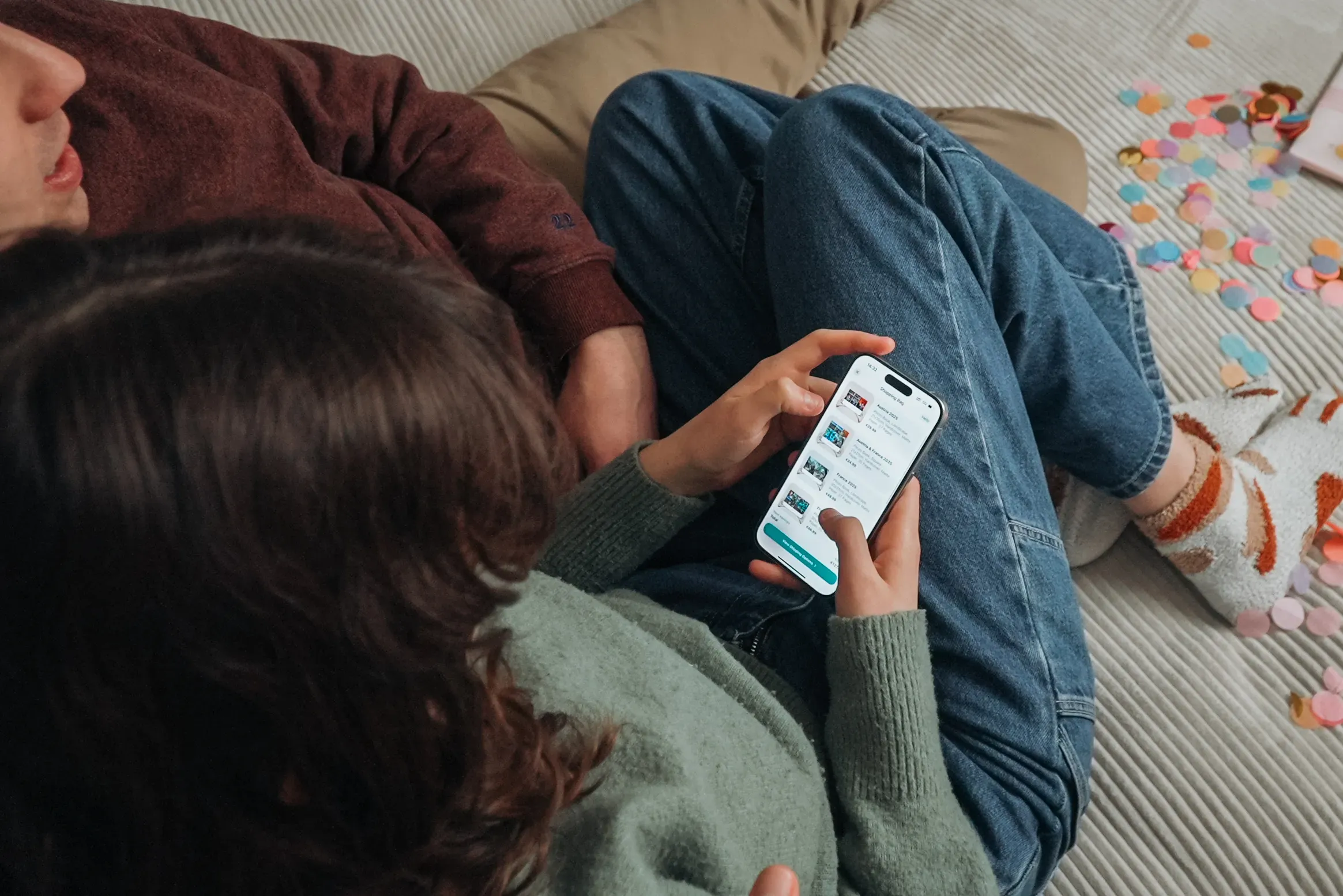 Two people sitting on a bed, one viewing a smartphone displaying a photo book making app. Colorful paper scraps scattered around.