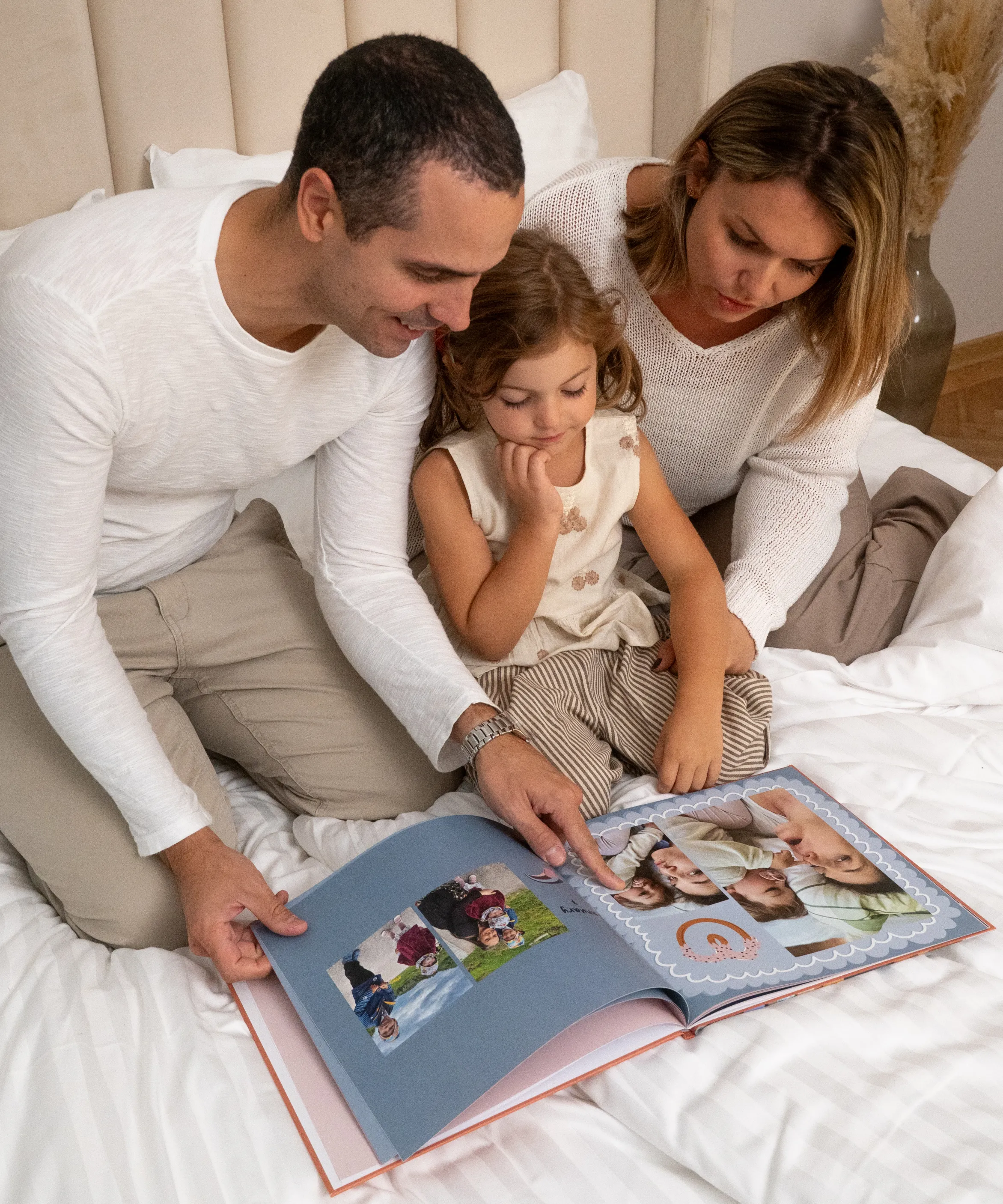 A family sitting on a bed, looking through a photo album together, with the little girl in the middle focused on the pictures.