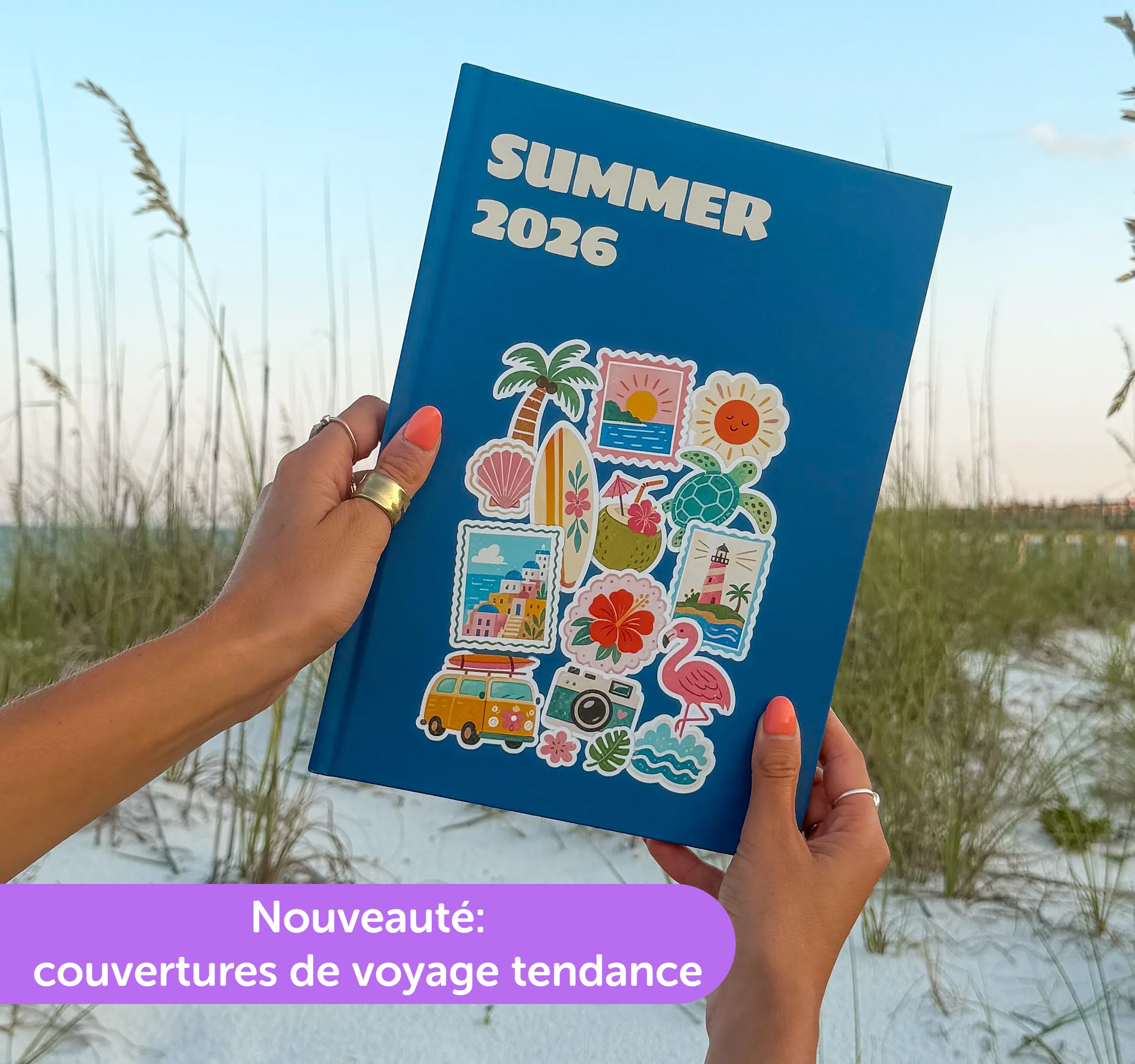 A person holds a blue journal labeled "Summer 2026" with vibrant travel stickers, set against a beach background with sand and grass.