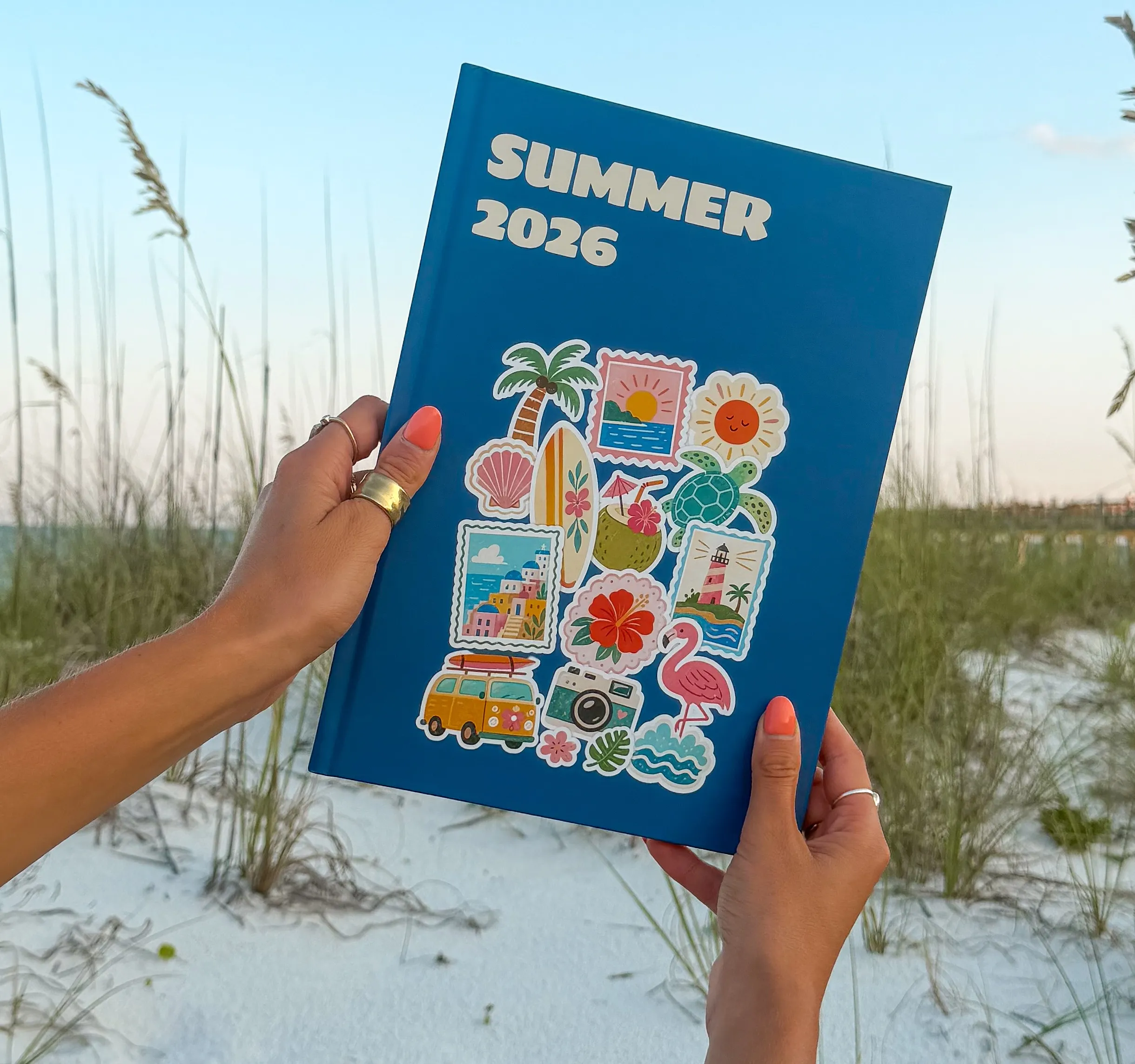 Person holding a blue notebook labeled "Summer 2026," decorated with colorful summer-themed stickers, with a sandy beach and grass in the background.