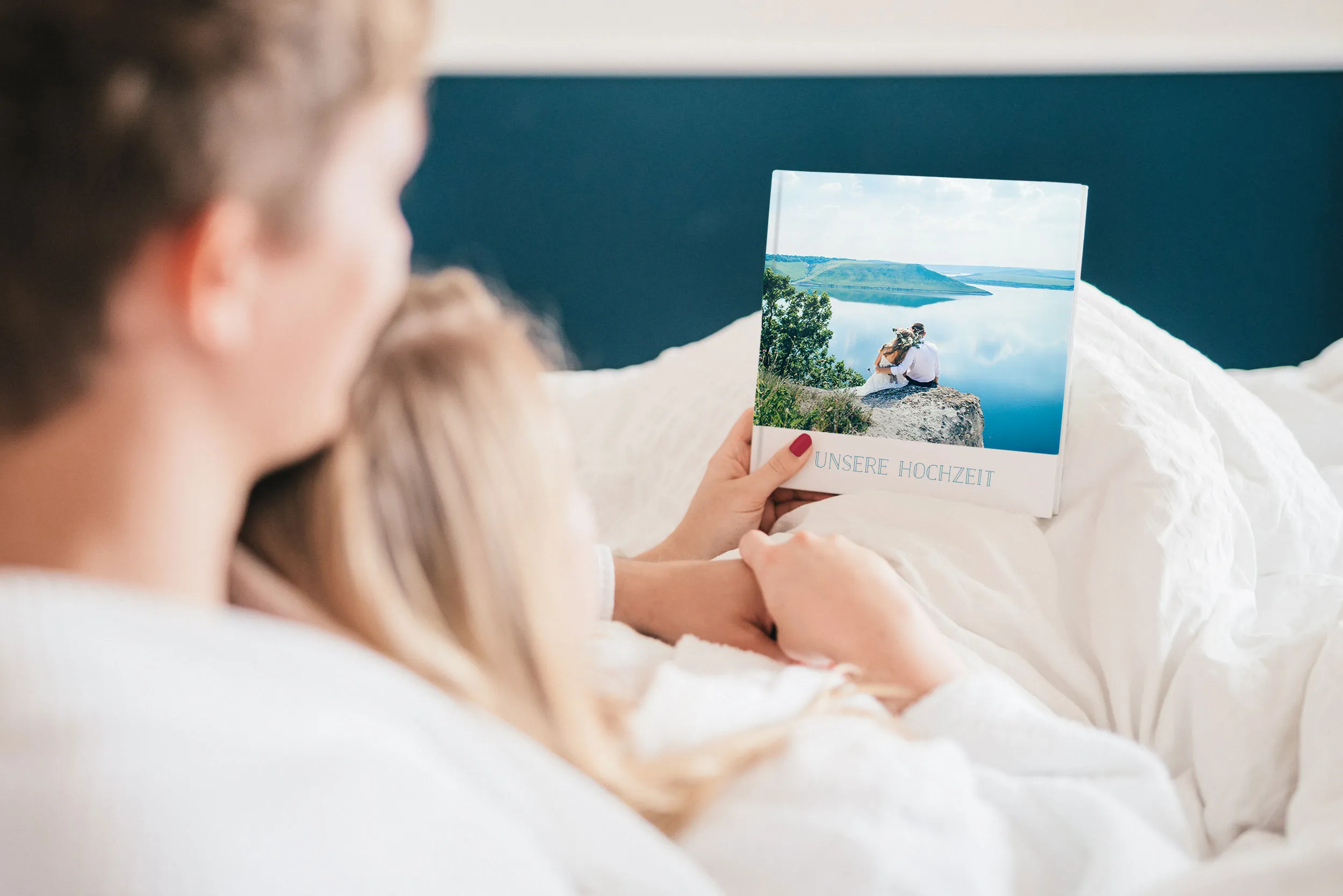 Ein Paar in weißen Gewändern chillt im Bett und hat ein Fotoalbum mit einem schönen Bild von einem Paar an einem See auf dem Cover in der Hand.