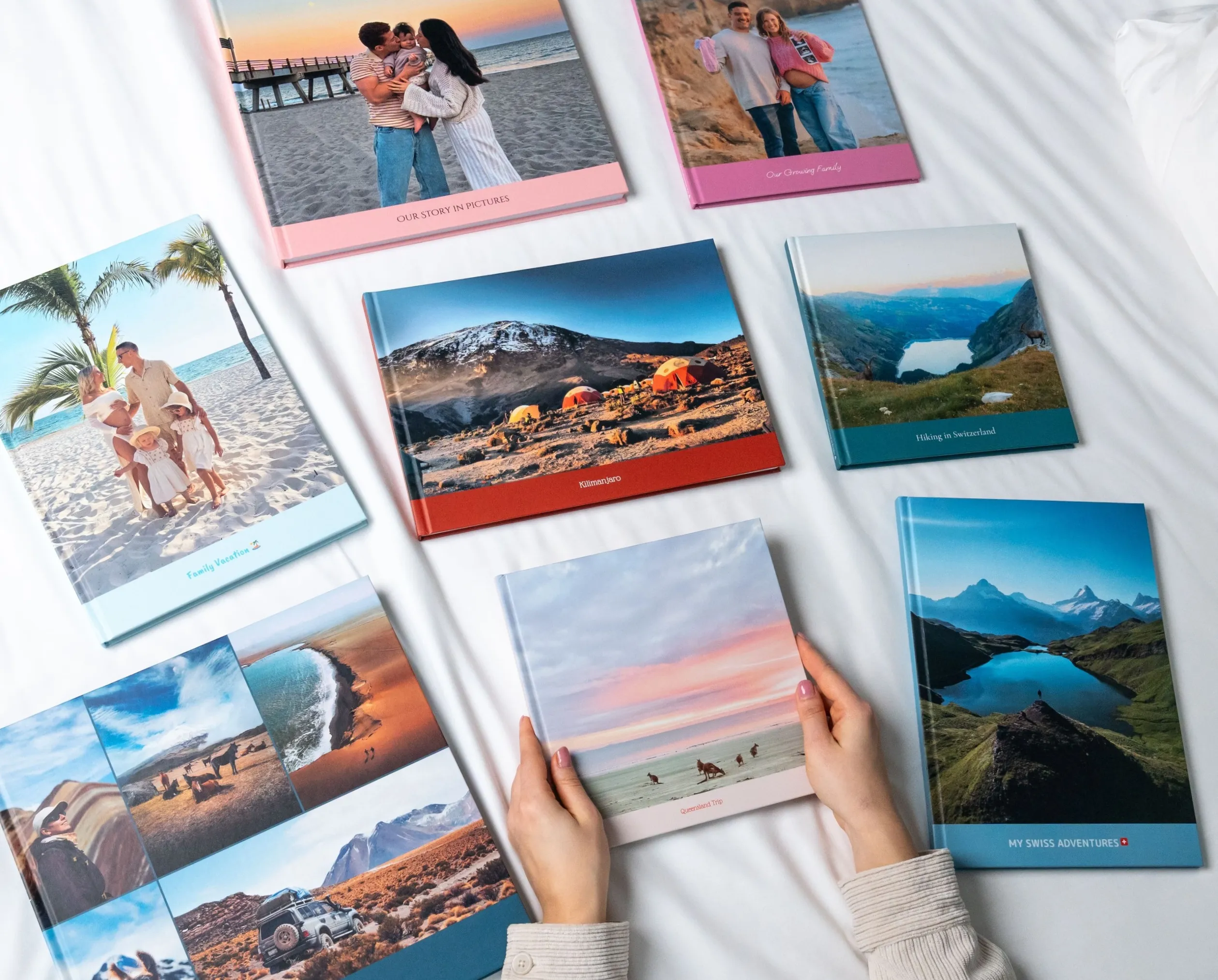 A person holds a photo book on a bed, surrounded by various colorful photo albums featuring travel and family pictures.