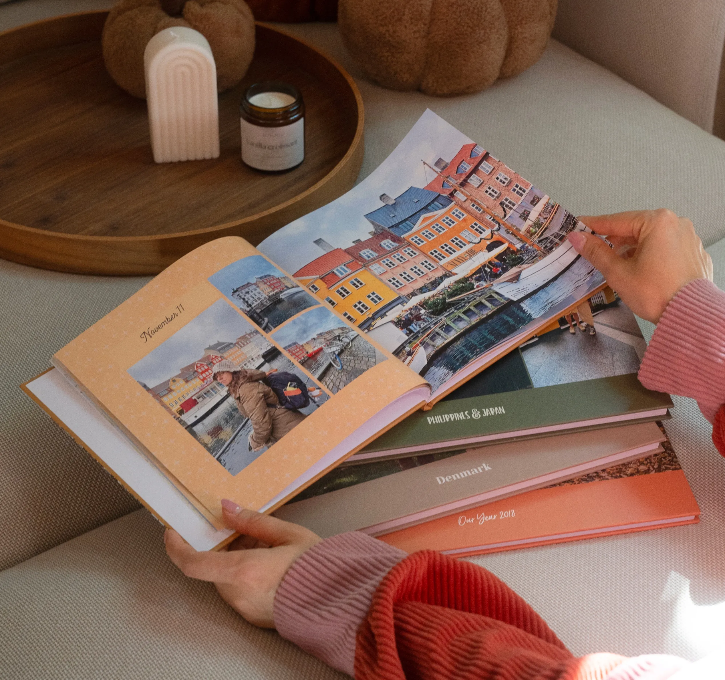 Hands holding a photo book open to images of colorful buildings by a canal, with stacks of other travel photo books nearby on a sofa.