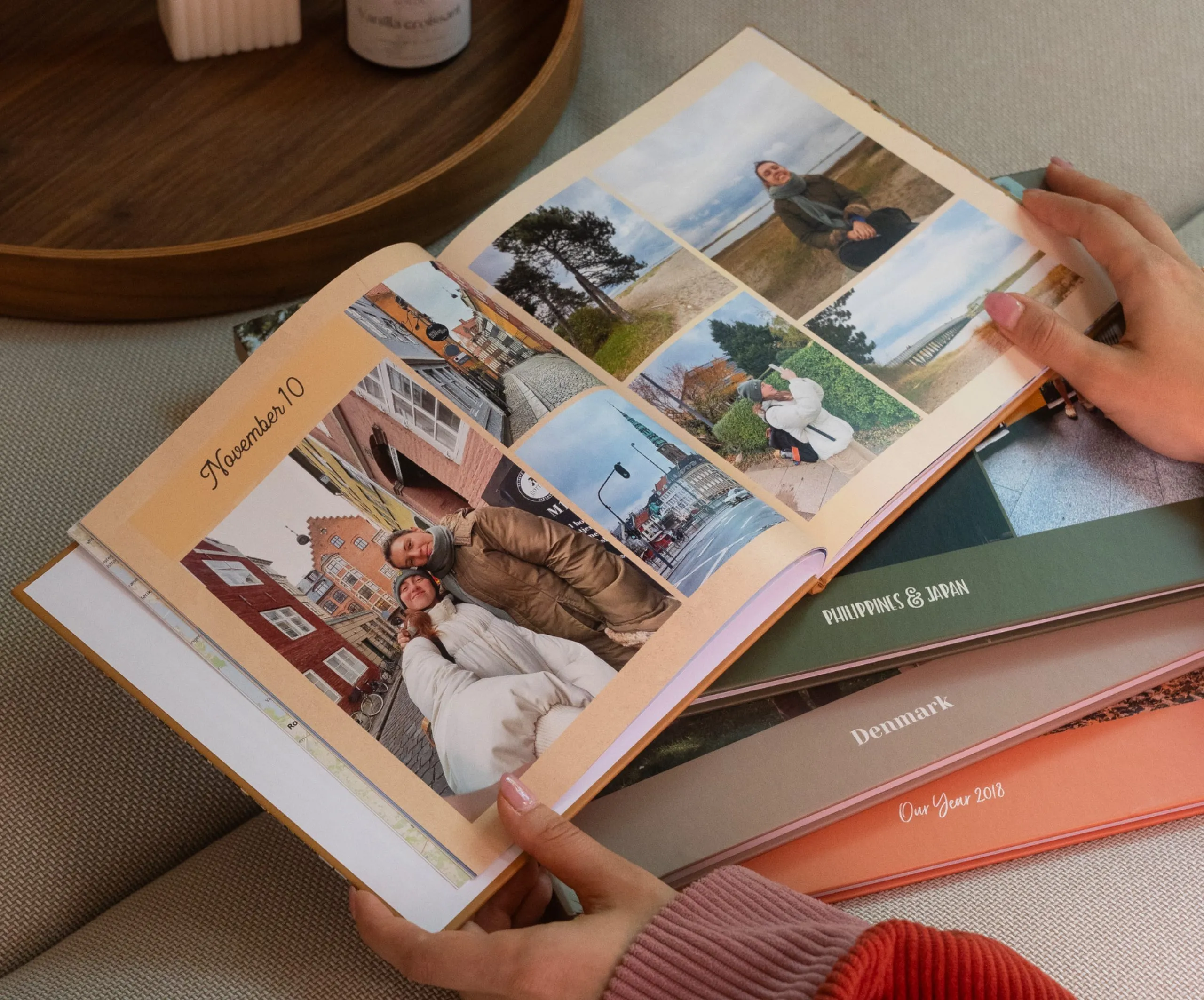 Hands holding an open photo book with travel pictures, surrounded by other closed books on a couch.