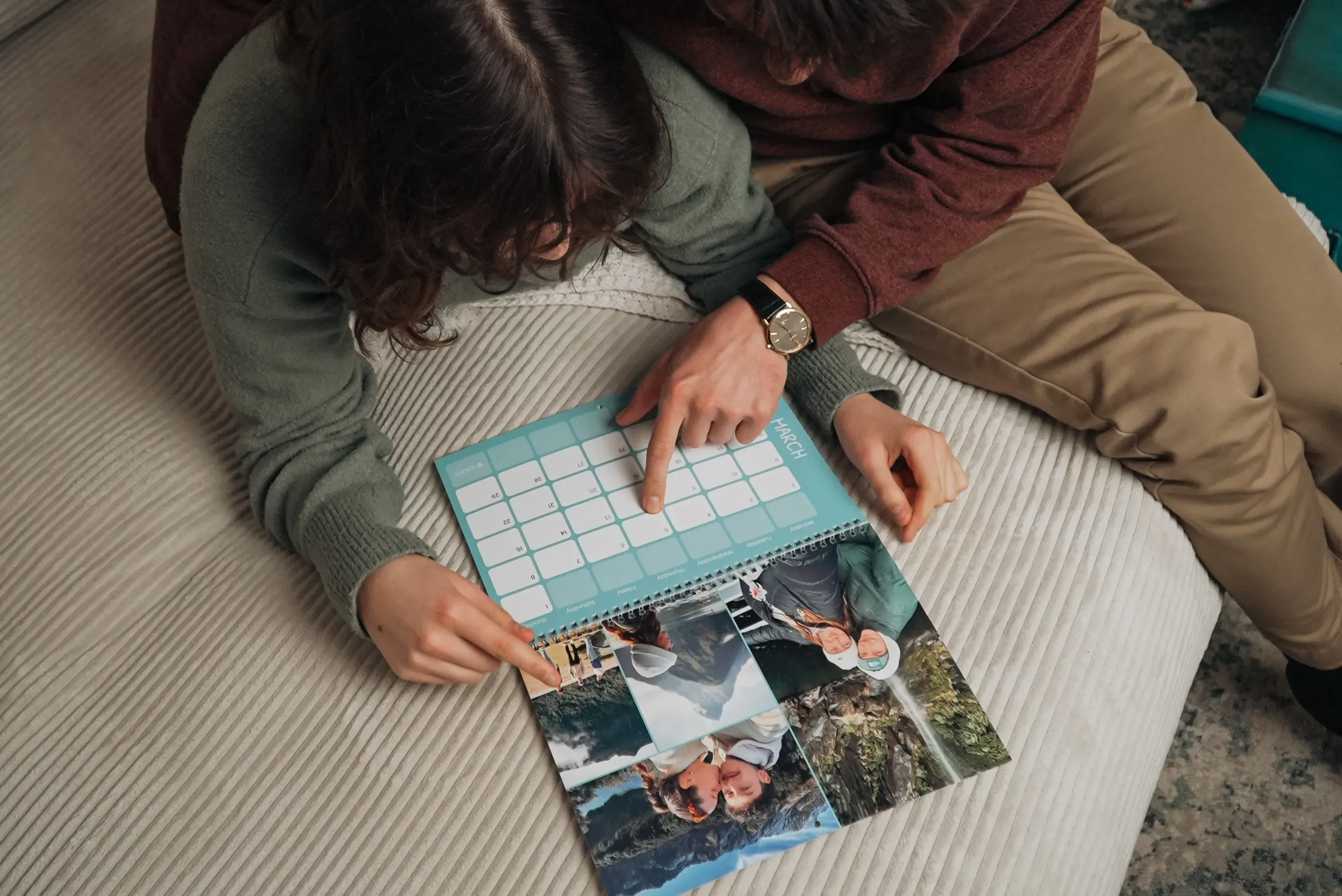 A couple sitting on a bed, looking at a calendar book with photos, while one person points at a specific date.