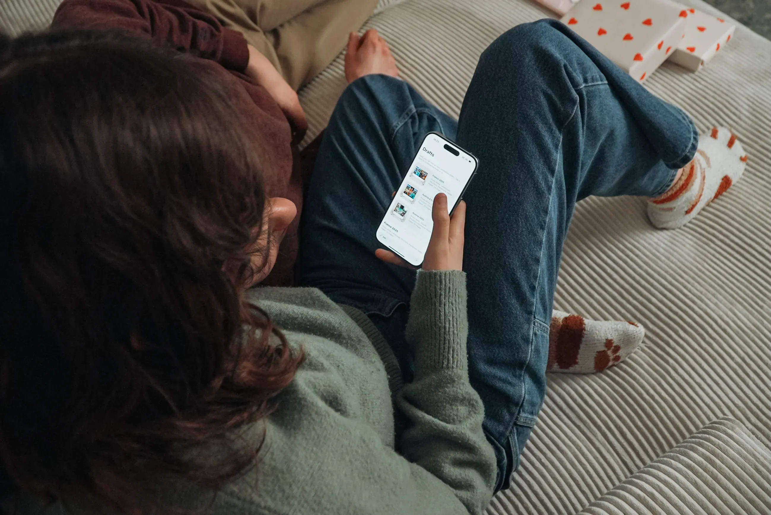 Person in a green sweater and jeans using a photo book app on their smartphone, sitting on a bed with a white blanket and patterned socks visible.