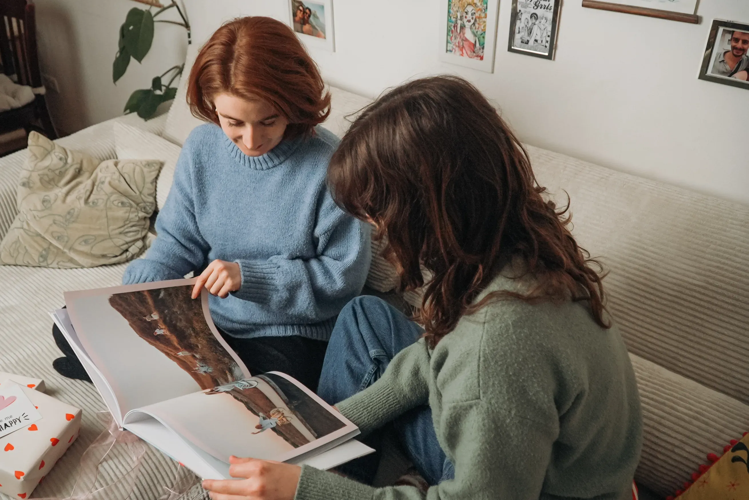 Two women sitting on a sofa, looking at a large open photo book with colorful images. The room has framed pictures on the wall.