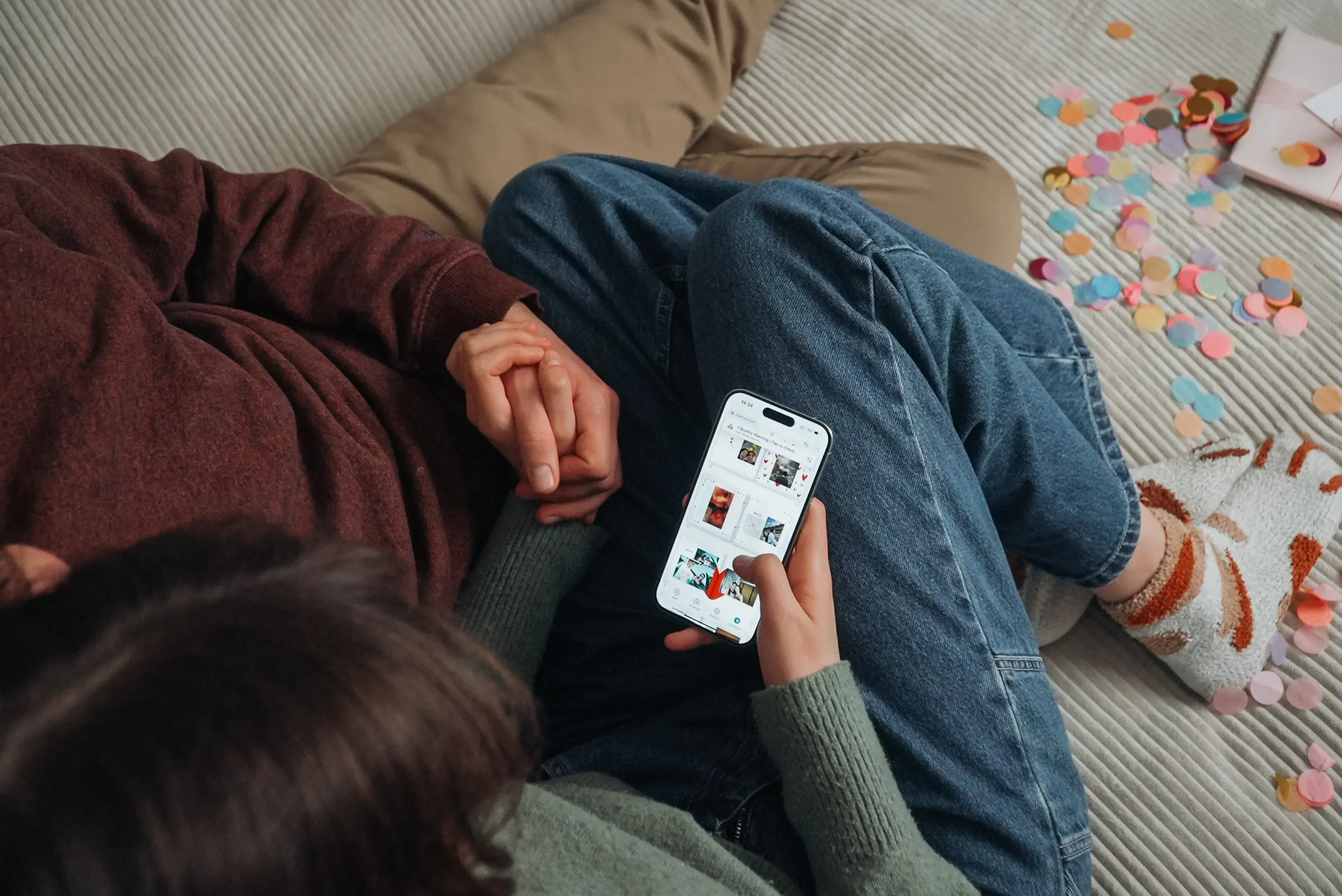 Two people sitting together, holding hands. One is browsing on a smartphone. They are surrounded by colorful paper hearts.
