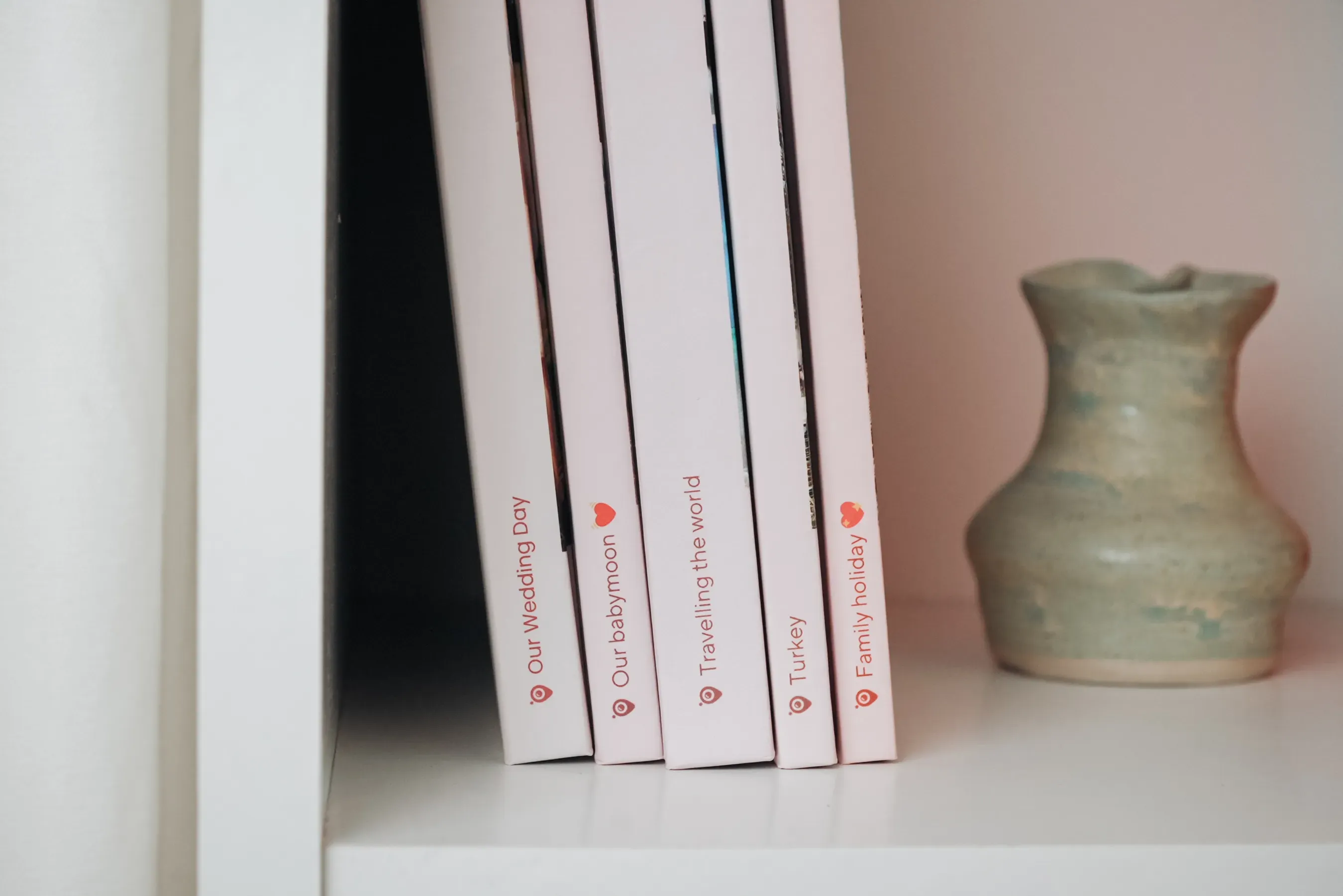photo Books with personal titles on a white shelf next to a small, light-colored ceramic vase.