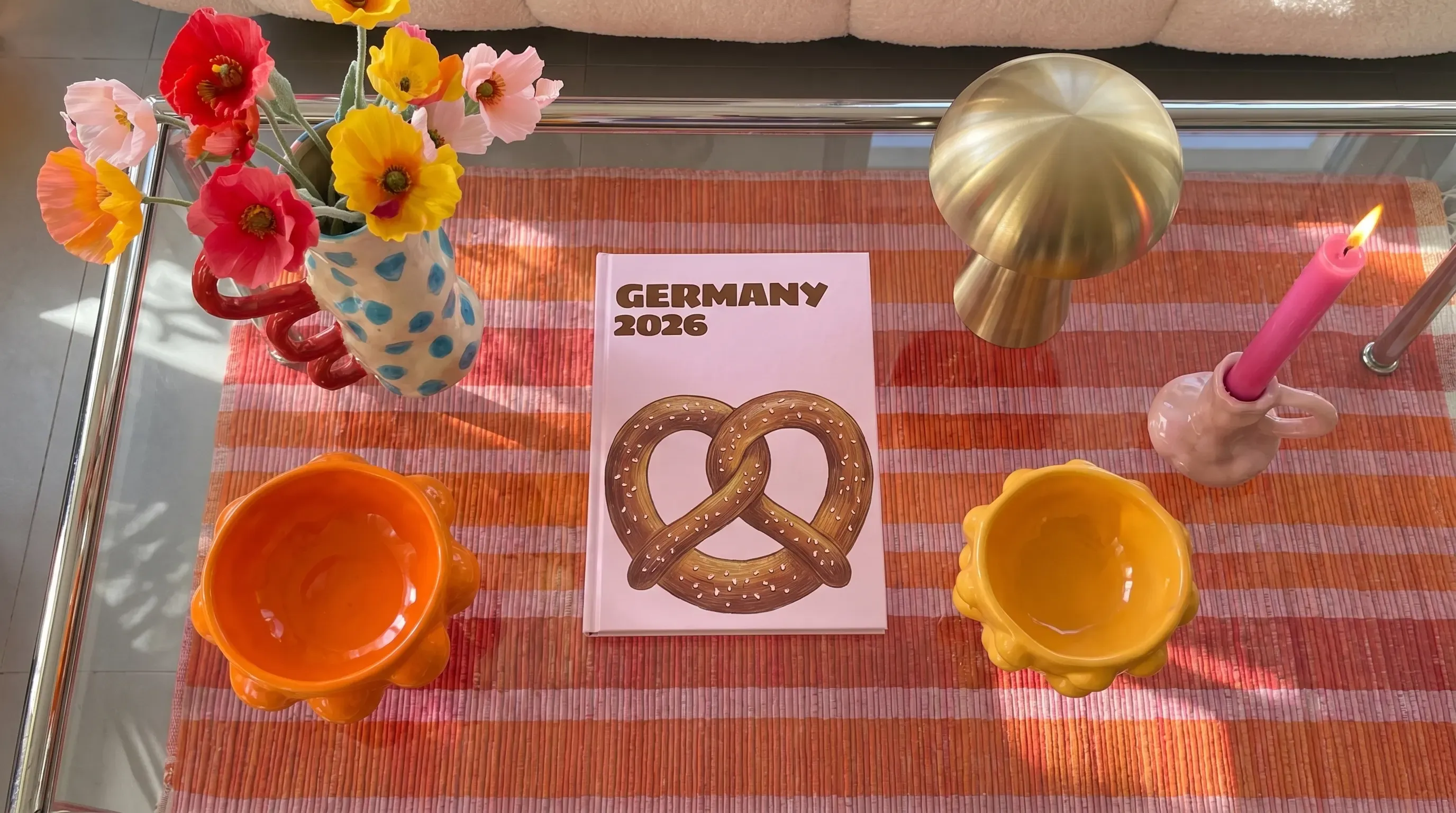 A glass coffee table with a Germany 2026 book featuring a pretzel, colorful flowers, vases, a brass lamp, candle, and orange and yellow bowls.