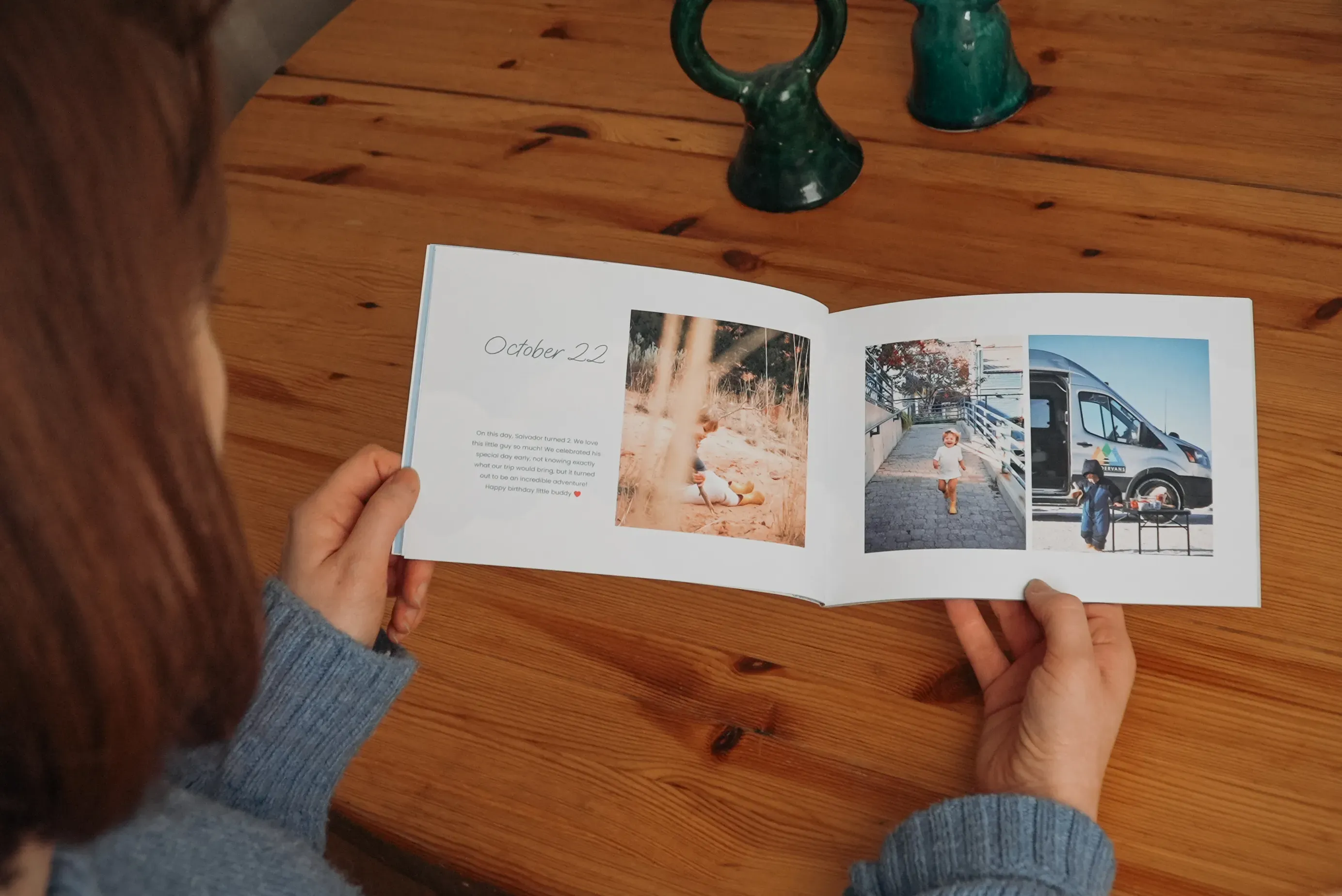 A person in a blue sweater holds an open photo book, displaying images and a date, "October 22," on a wooden table.