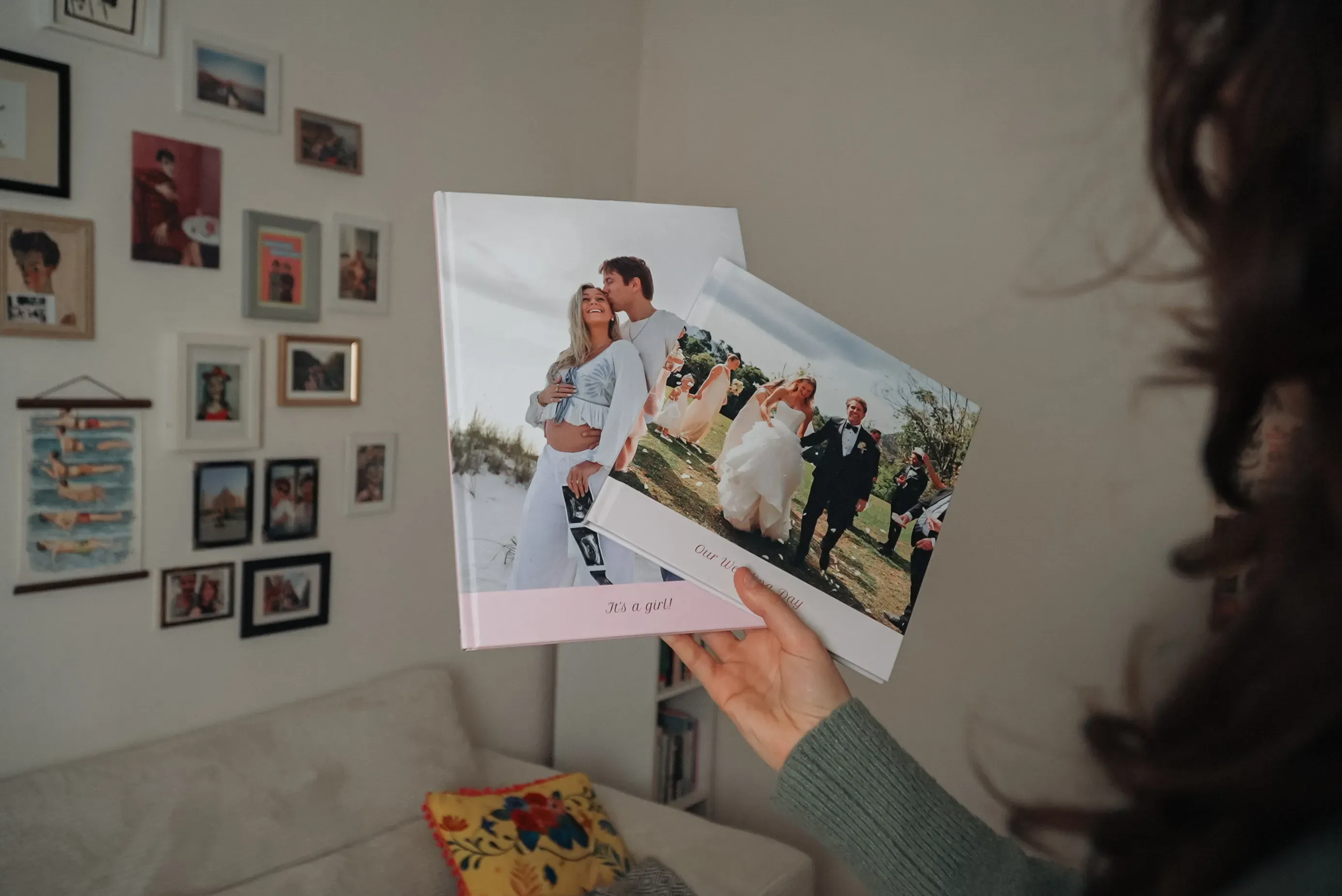 Hand holding two wedding photo albums, one with a kissing couple on the cover; wall decorated with framed pictures in the background.
