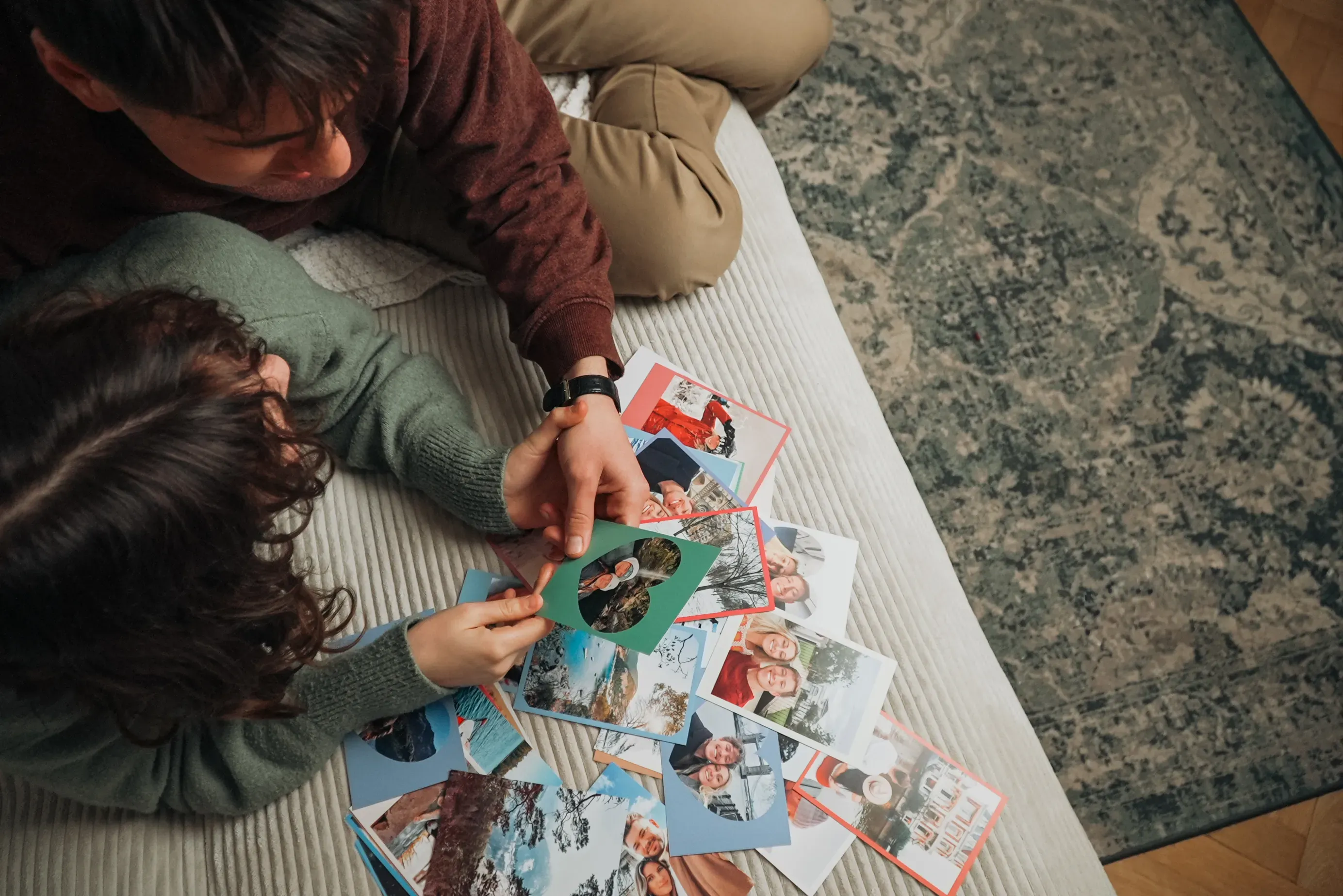 Two people looking at a collection of photo cards and photos on a bed. Some cards feature winter scenes and family pictures.
