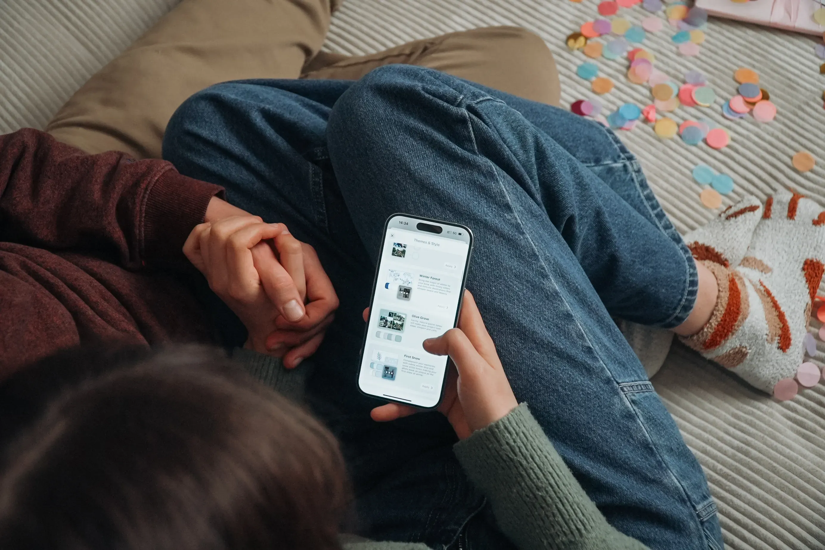 Eine Person sitzt mit gekreuzten Beinen da, hält die Hand einer anderen Person und scrollt durch die Journi Fotobuch App. Auf dem Boden liegen bunte Papierfiguren verstreut; man sieht gemütliche Socken.