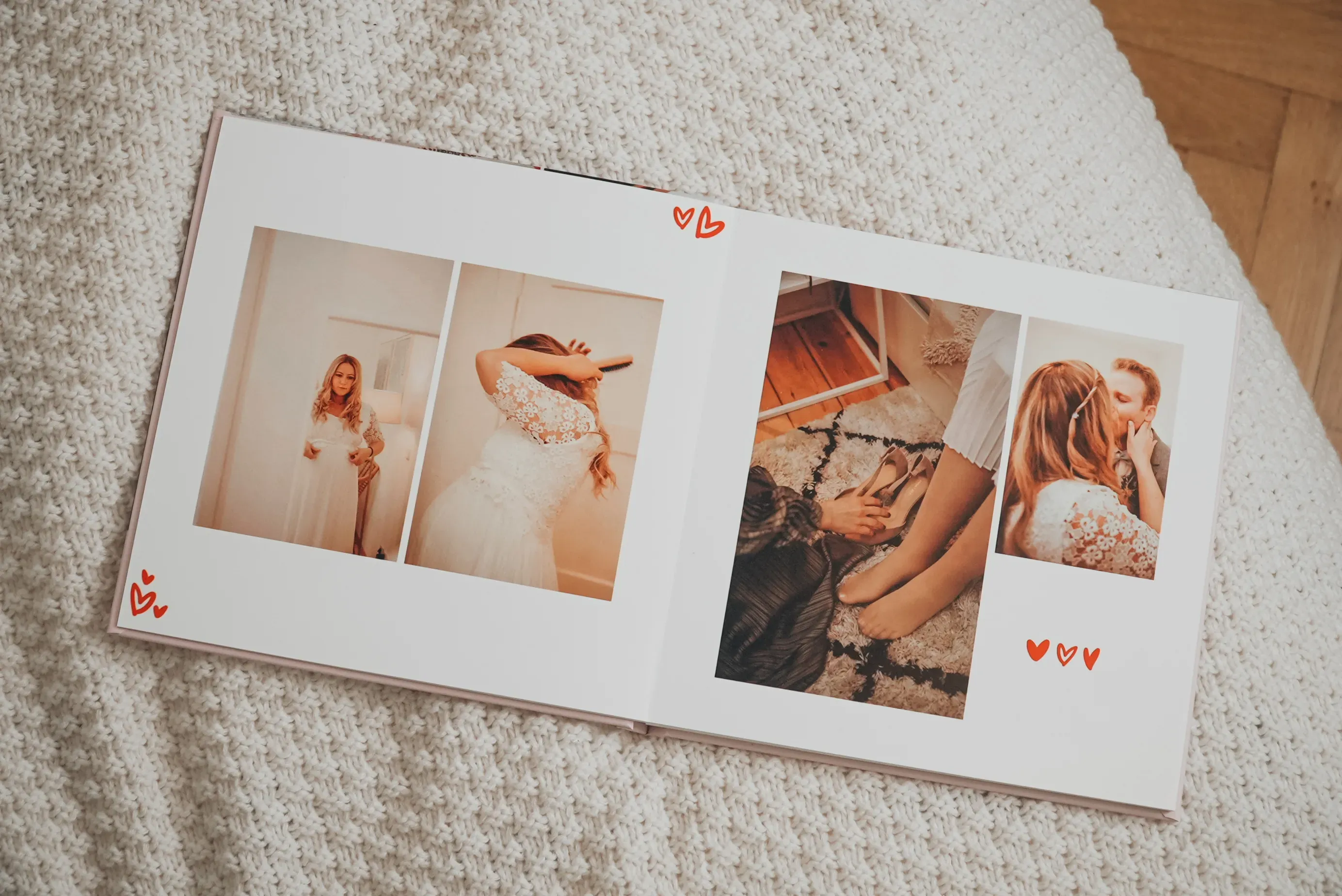 Offenes Fotoalbum auf einer strukturierten Oberfläche, das eine Braut in einem weißen Kleid zeigt, die sich fertig macht und beim Anziehen ihrer Schuhe geholfen bekommt; rote Herzen schmücken die Seiten.