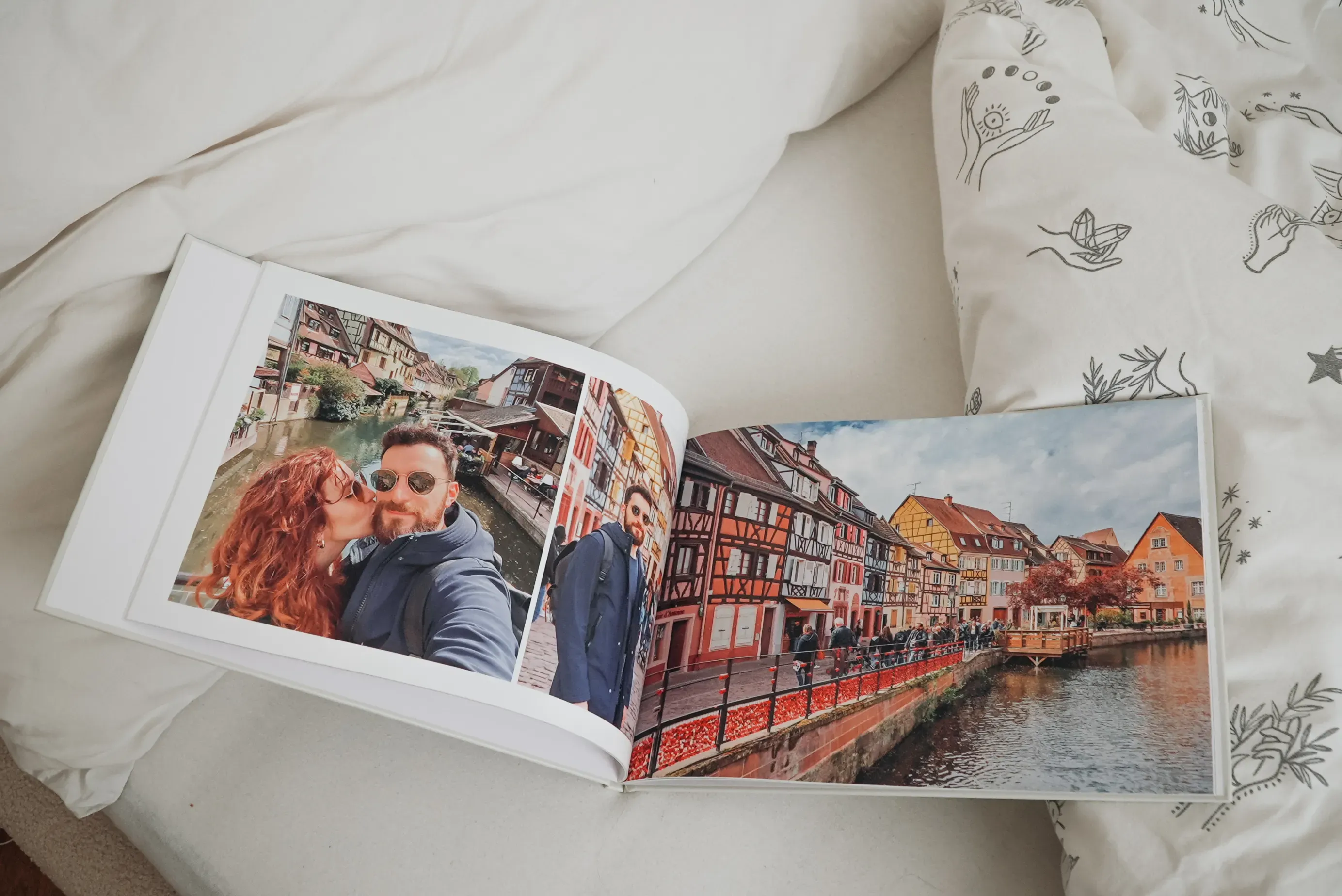 Open photo book on a bed, featuring a couple's travel photos in a picturesque European town with colorful buildings by a canal.