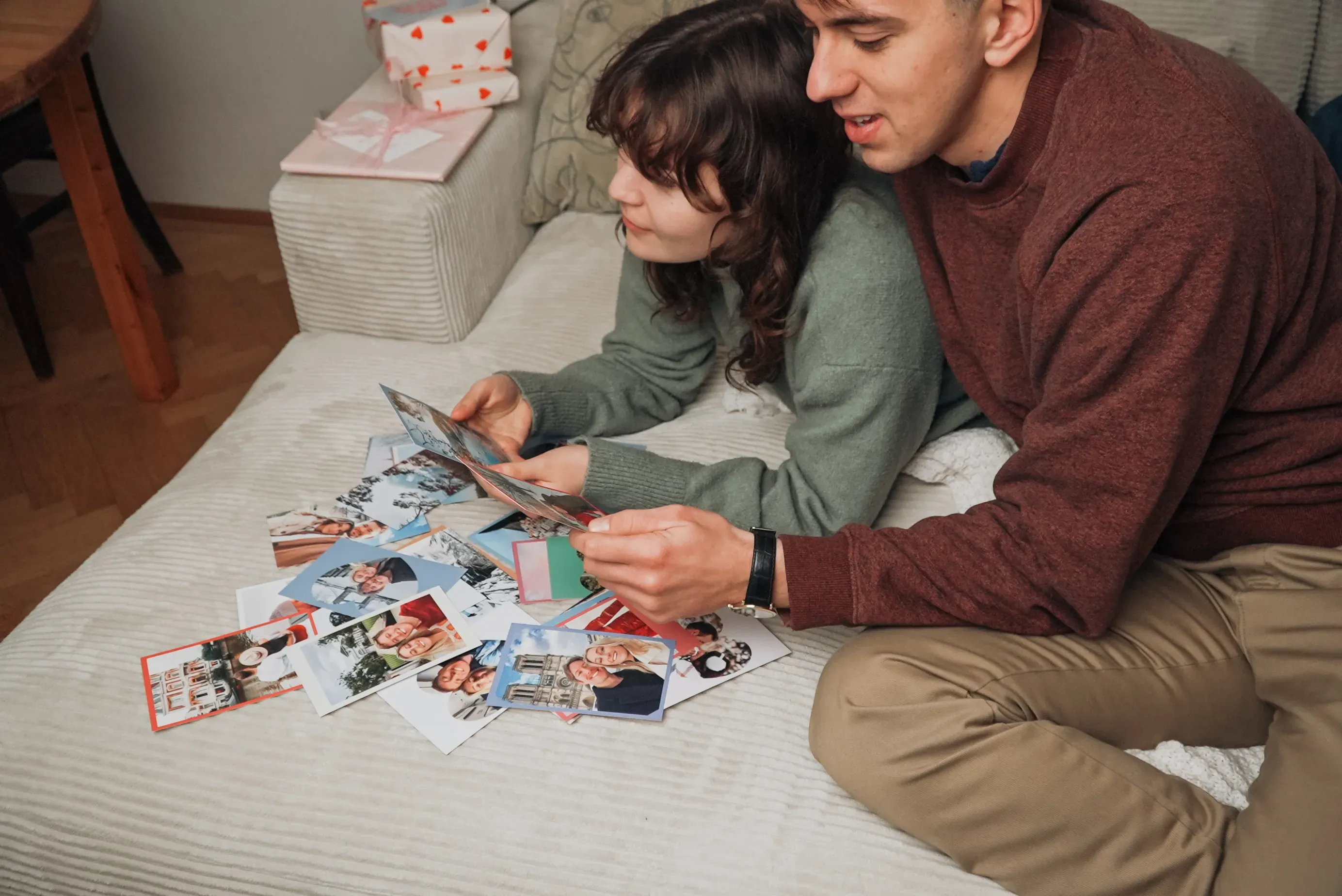 A couple lounges on a couch, looking at printed photo cards spread out in front of them, with wrapped gifts in the background.