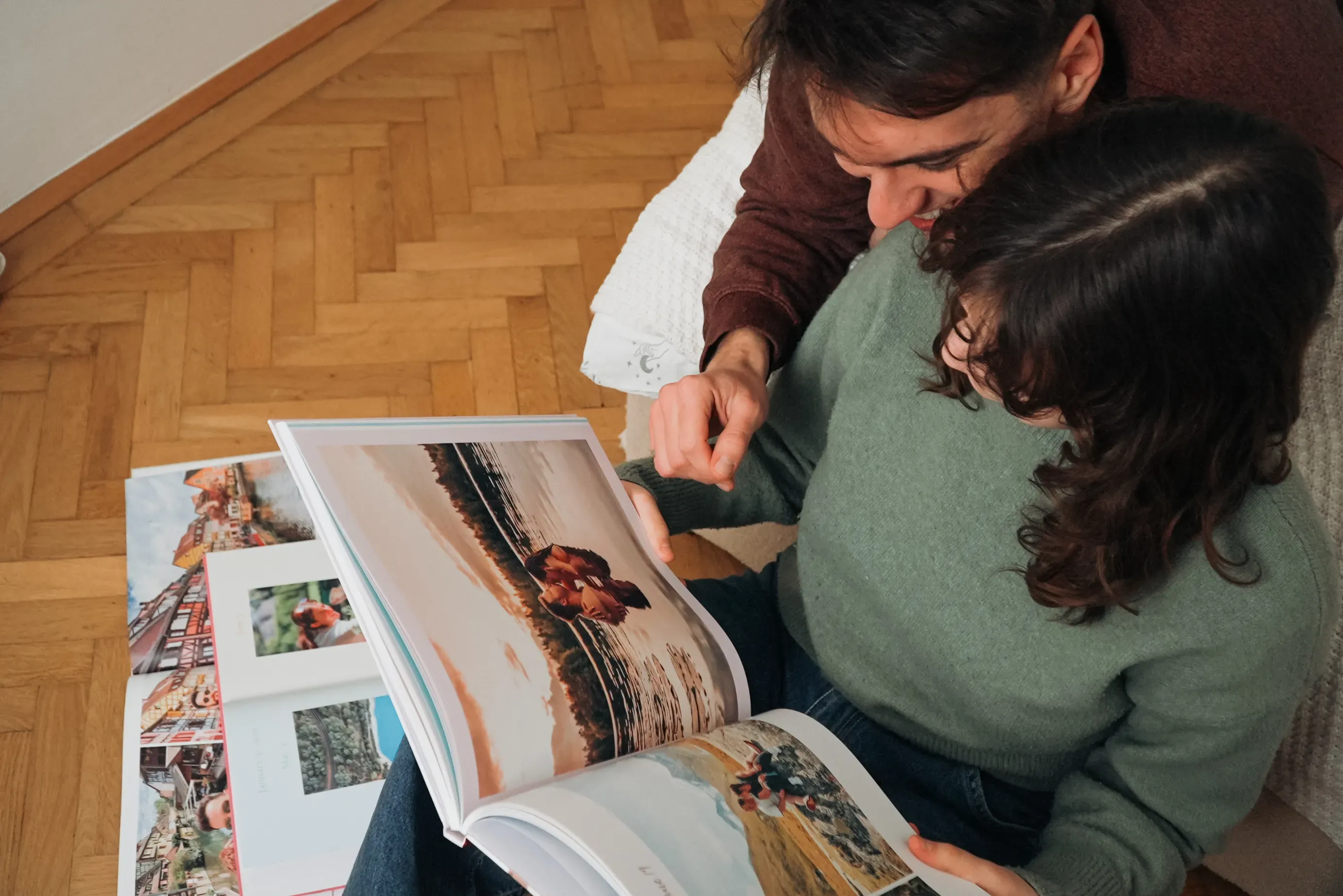 A couple sits on the floor, looking at a an anniversary photo book filled with travel pictures, in a cozy room with wooden flooring.