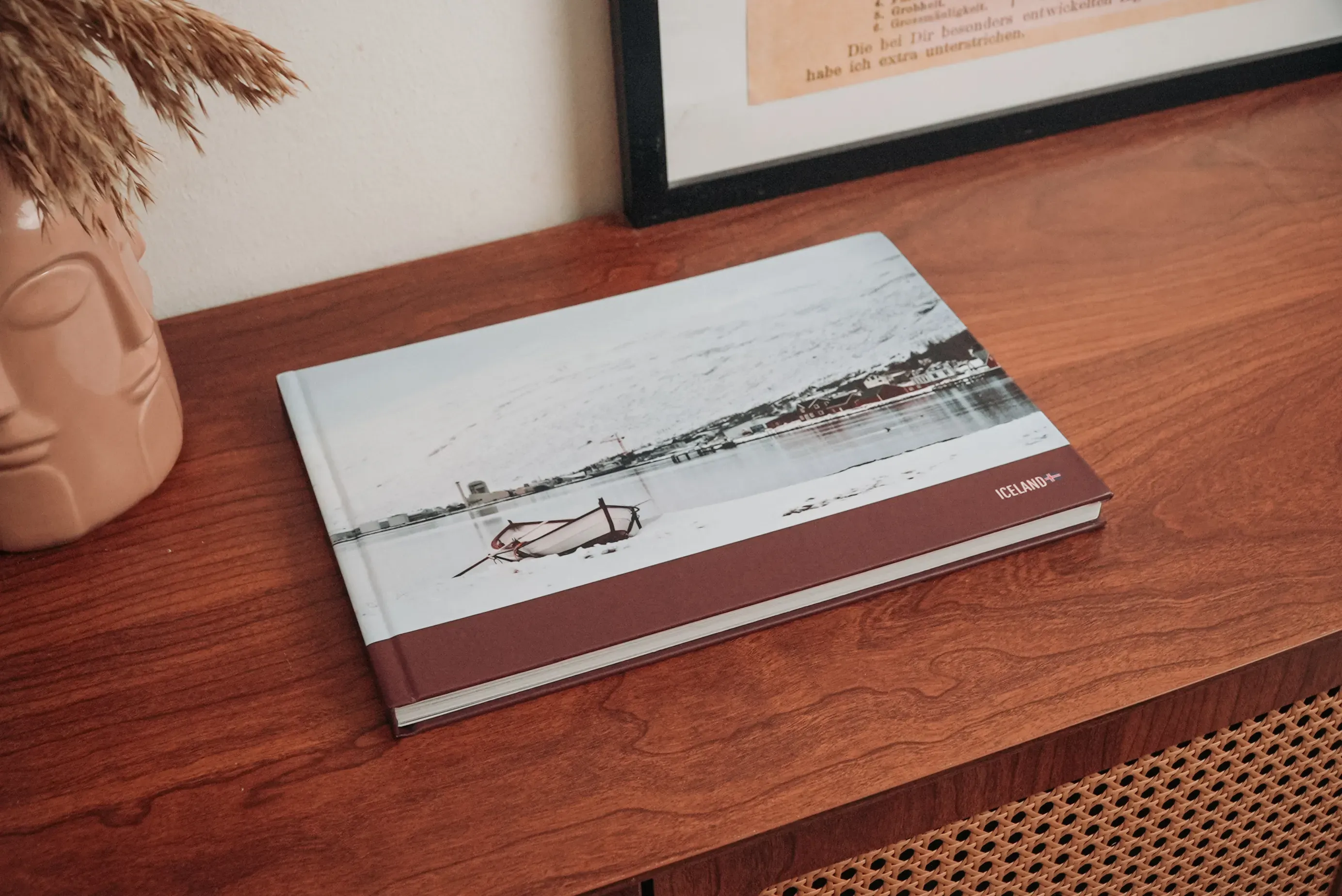 A hardcover photo book with a snowy landscape cover rests on a wooden coffee table near a vase with dried plants and a framed picture.