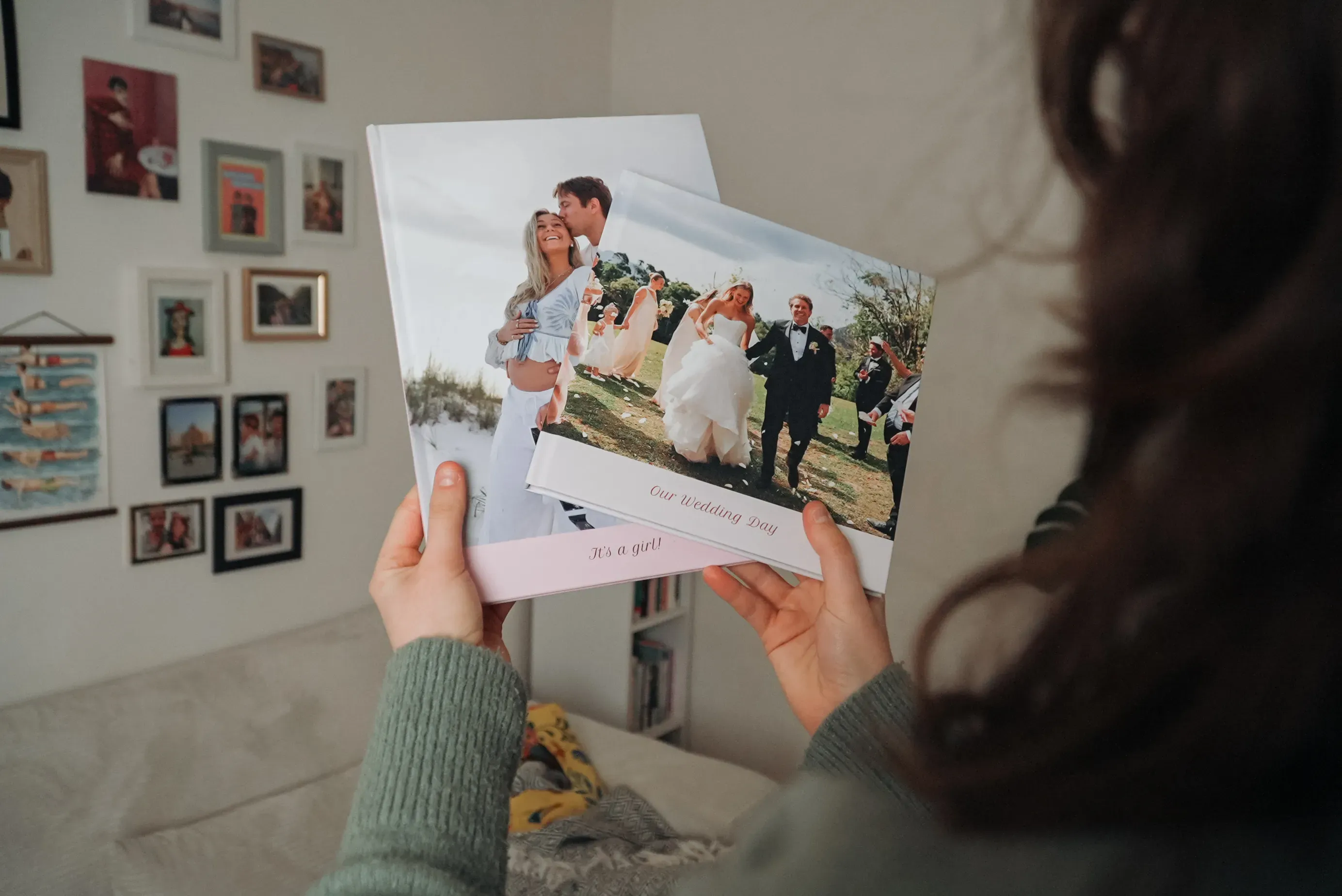 Person holding two wedding photo albums, with wall photos in the background, in a cozy room setting.