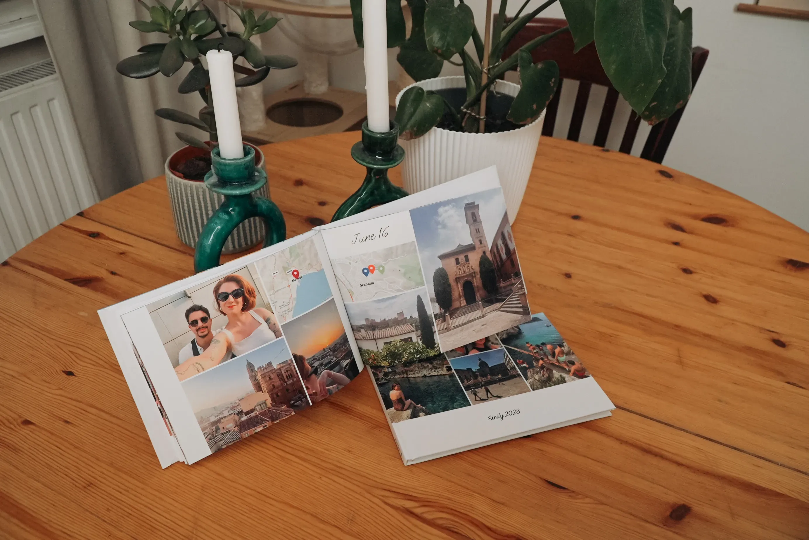 Two open photo books on a wooden table, displaying travel photos and dates. Green candle holders and plants are in the background.