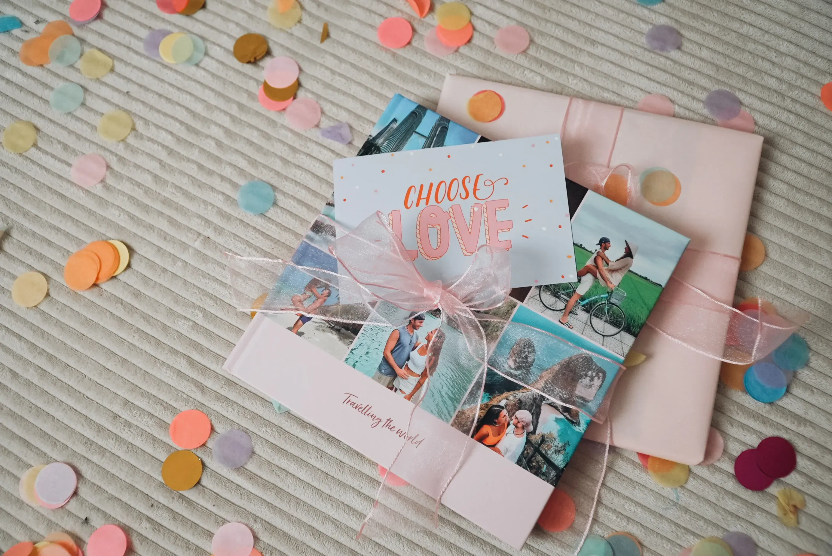 Gift-wrapped photo book with travel photos, a "Choose Love" card, and pastel confetti scattered on a textured surface.
