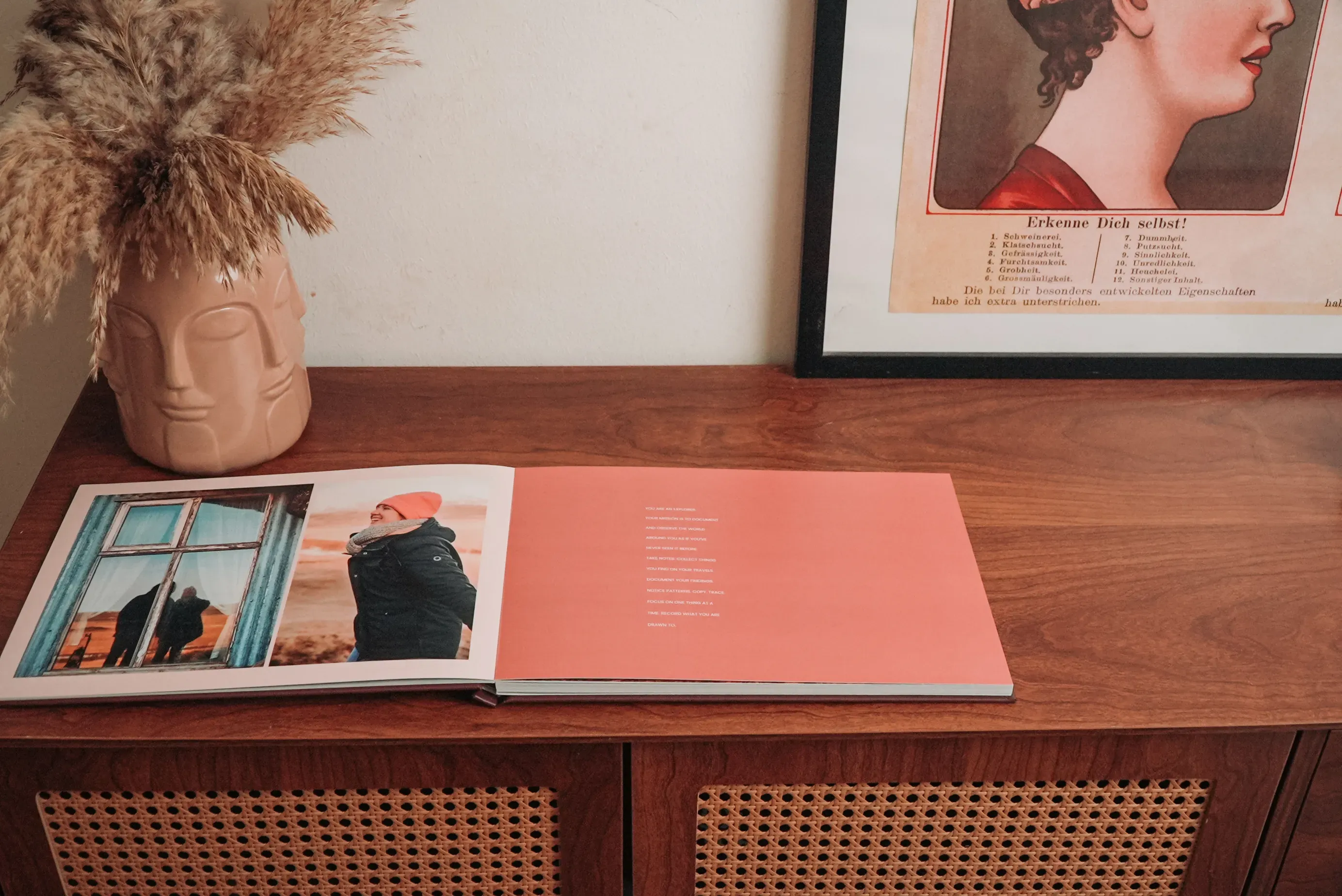 Open layflat  book on a wooden sideboard, next to a vase with dried flowers and a framed artwork. The book shows a photo of a person by a window.