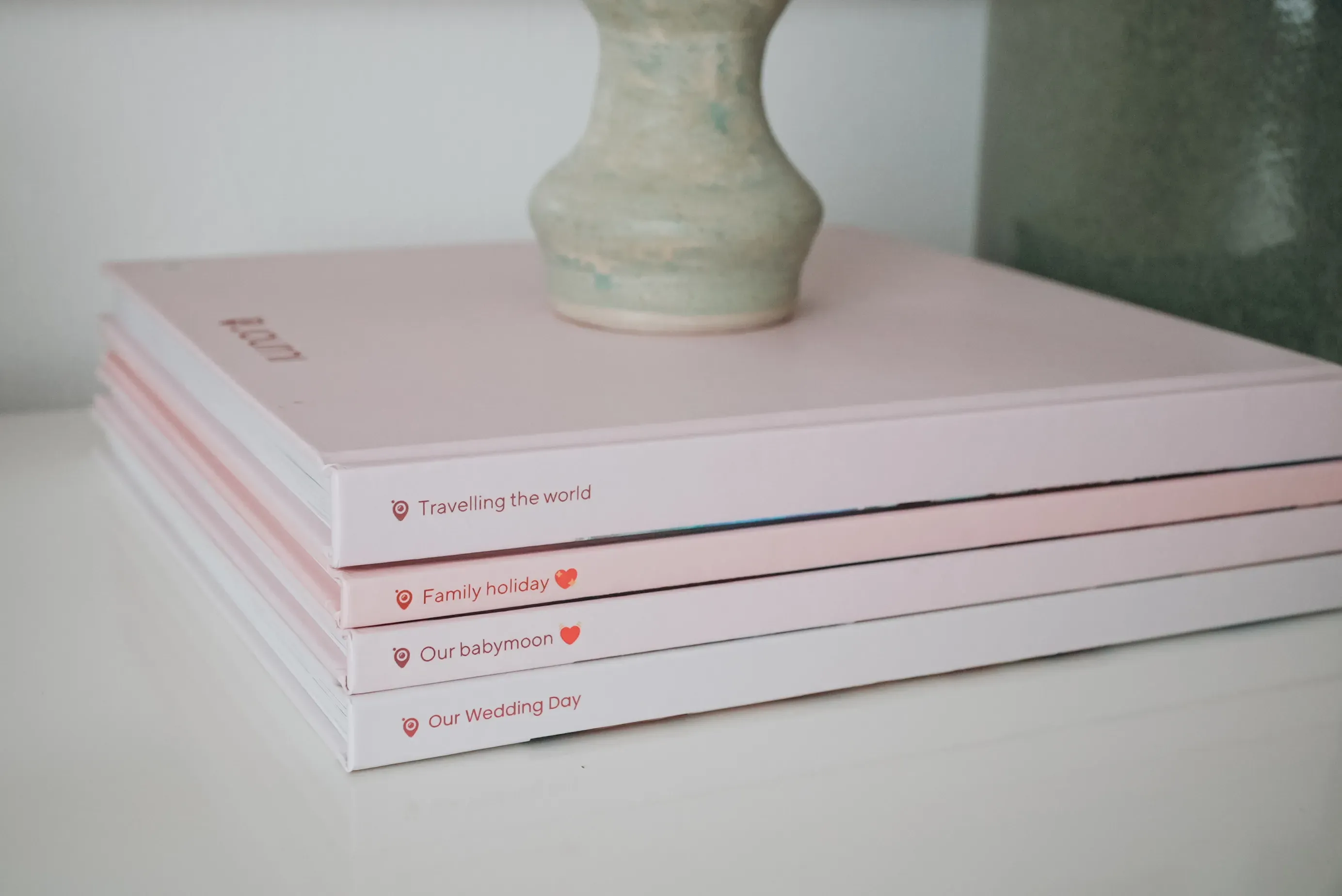 A stack of pink photo albums, each labeled with heart symbols: "Travelling the world," "Family holiday," "Our babymoon," "Our Wedding Day."